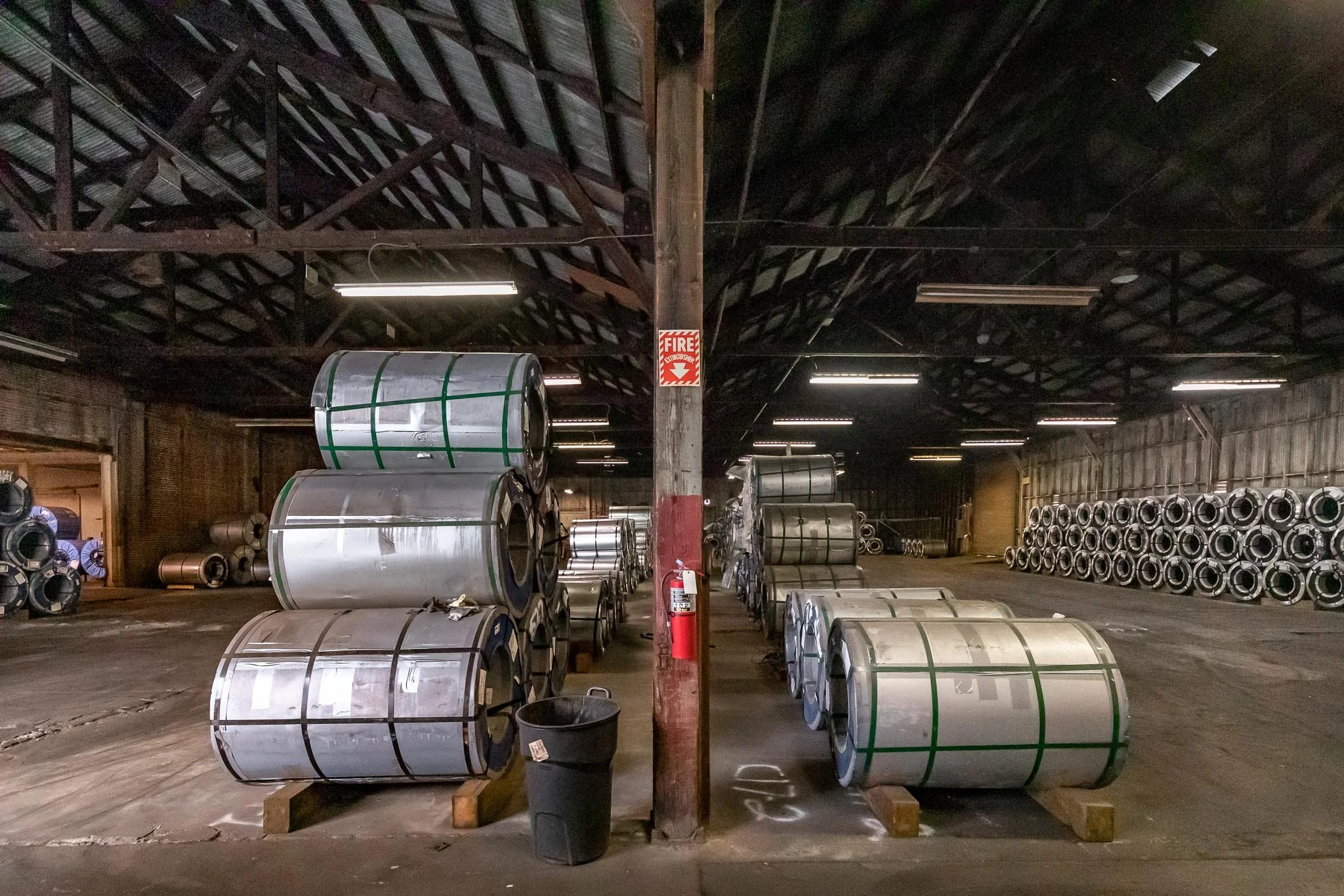 Large rolls of metal on wooden supports inside an industrial warehouse with a high wooden ceiling, brick walls, and fluorescent lighting.