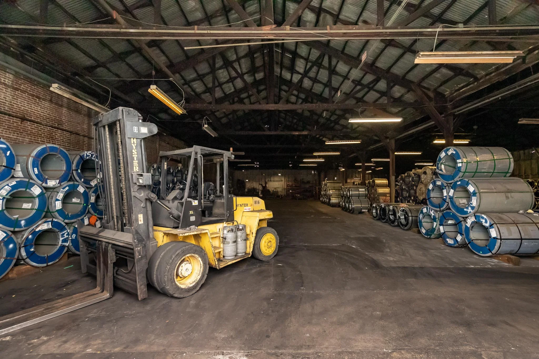 Warehouse with stacks of metal pipes on both sides and a yellow forklift in the center.