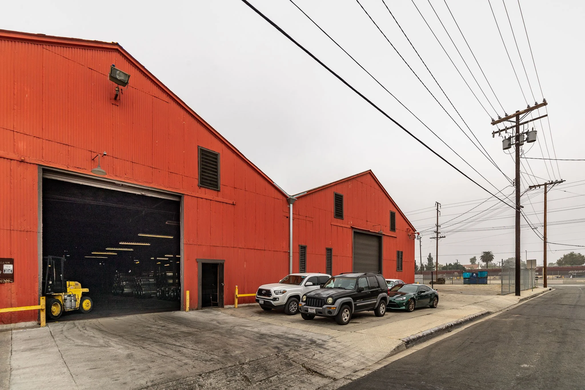 Red industrial warehouse with open large garage door, parked black, white, and green cars in front, utility poles and power lines in the background on an overcast day.