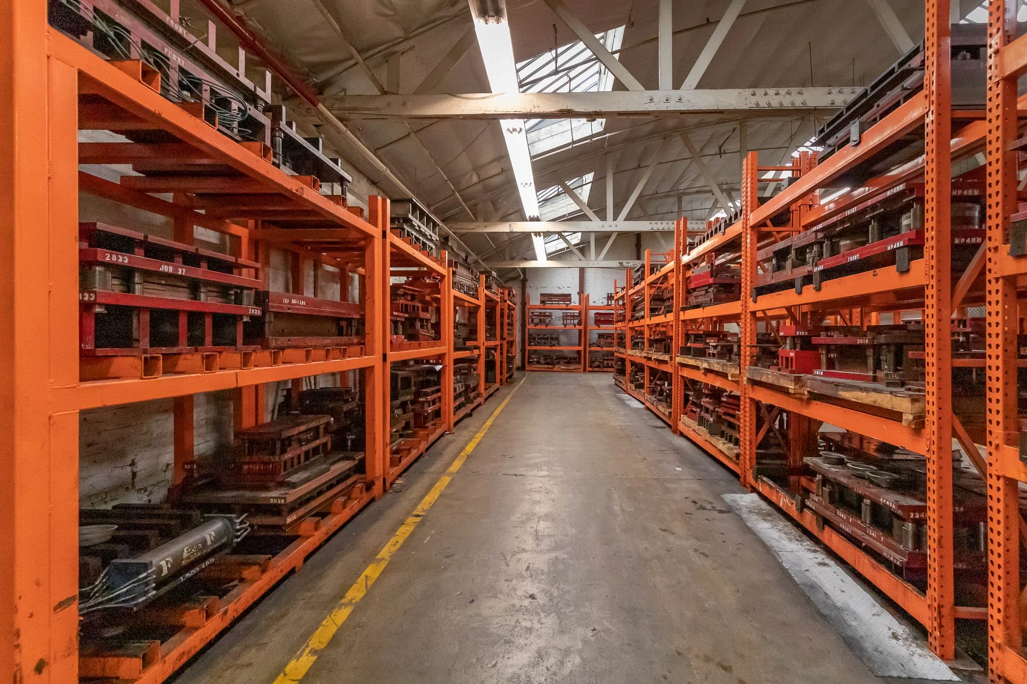 Warehouse aisle with orange metal shelves stocked with metal parts and tools under a high ceiling with skylights.