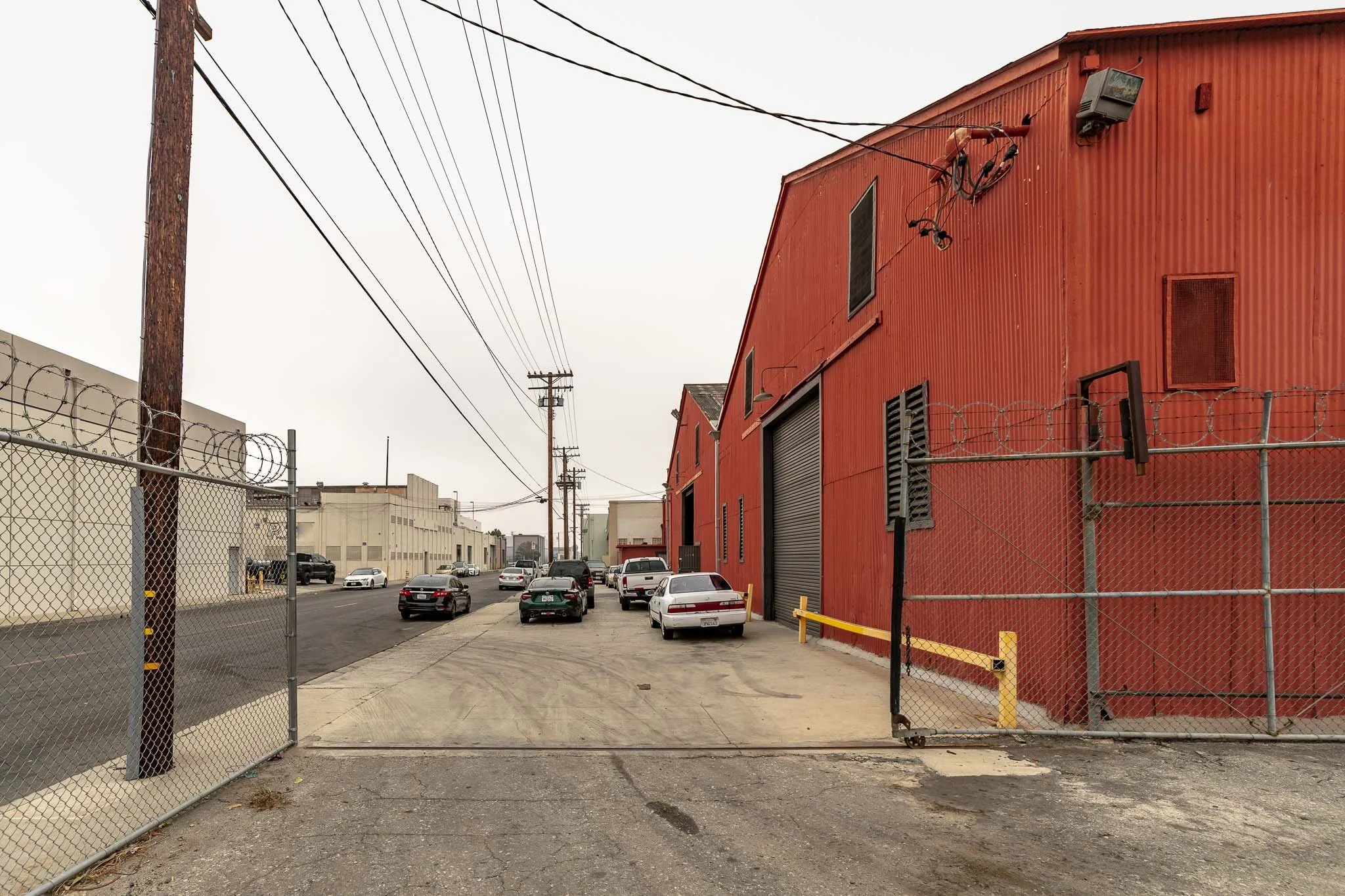 An industrial alley with a red warehouse on the right, chain-link fences, parked cars, utility poles, and power lines under an overcast sky.