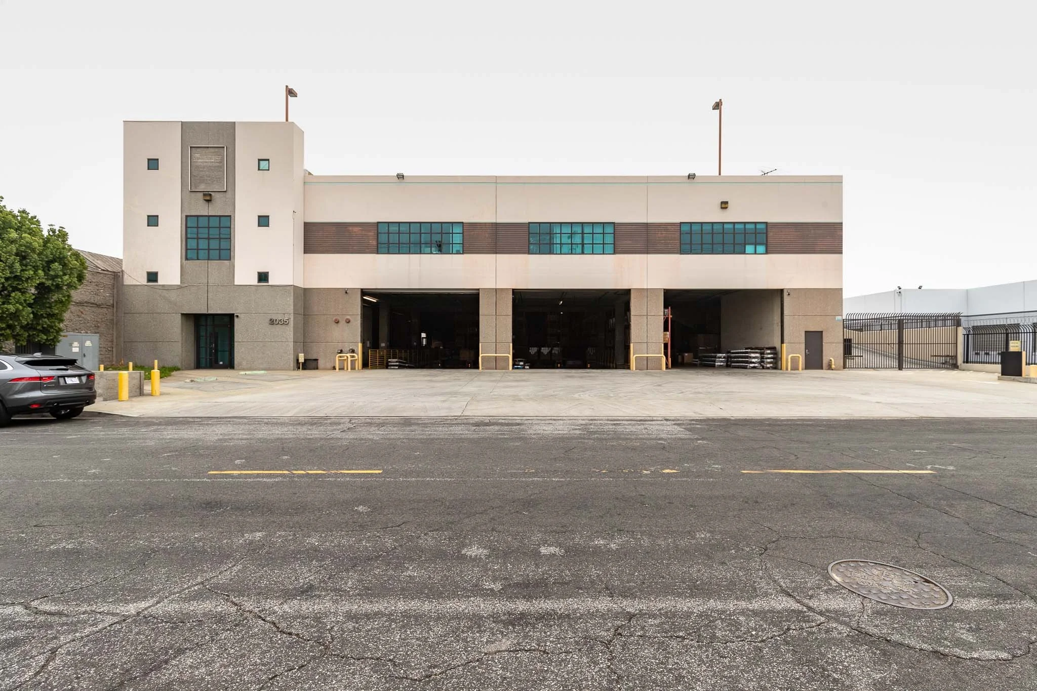 Large industrial warehouse building with open loading bays, parked pallet stacks, a black car in front, and a paved street with yellow markers.