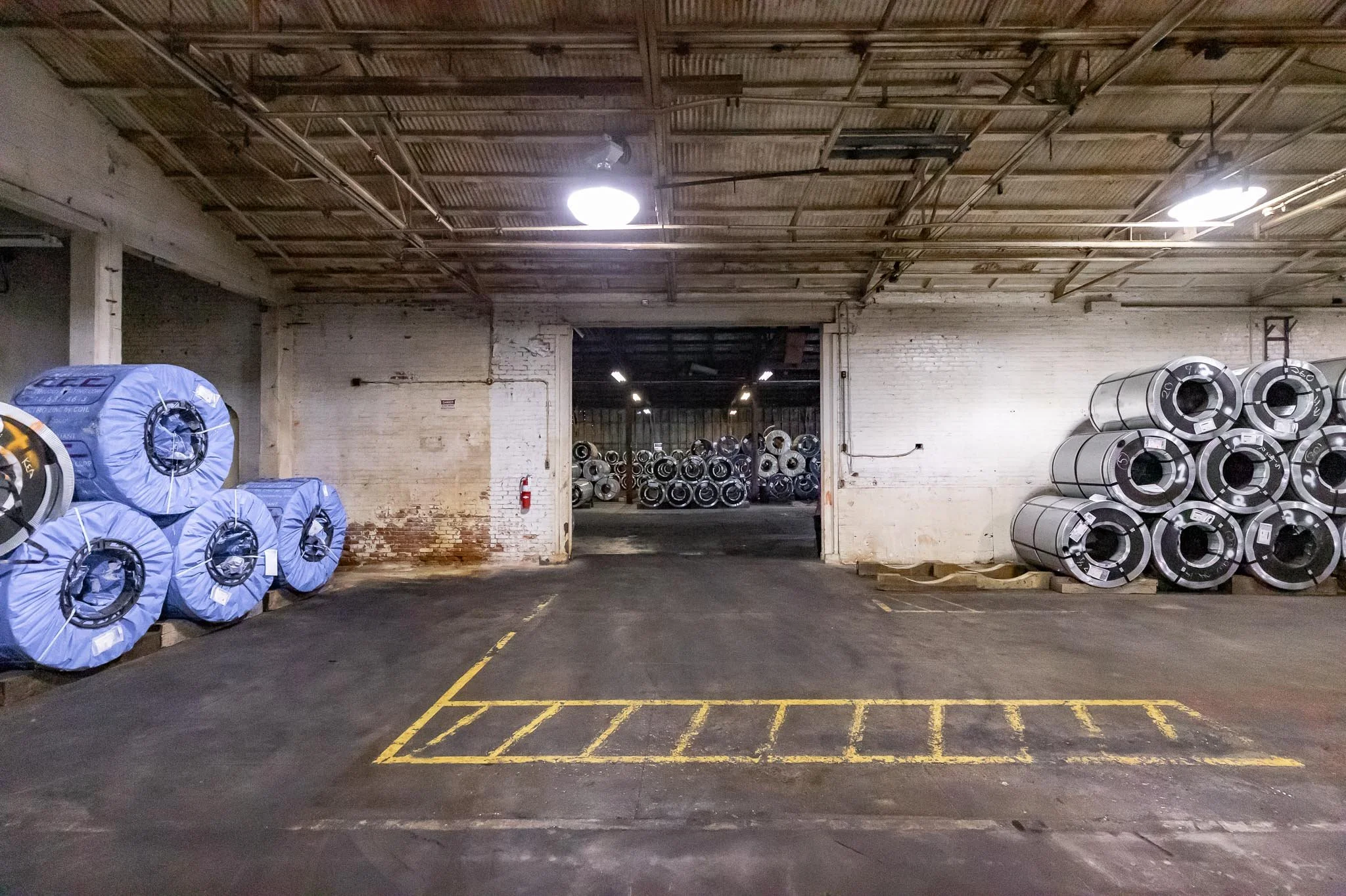 Indoor warehouse with large rolls of metal on each side and a partially open doorway leading to more metal rolls in the background.