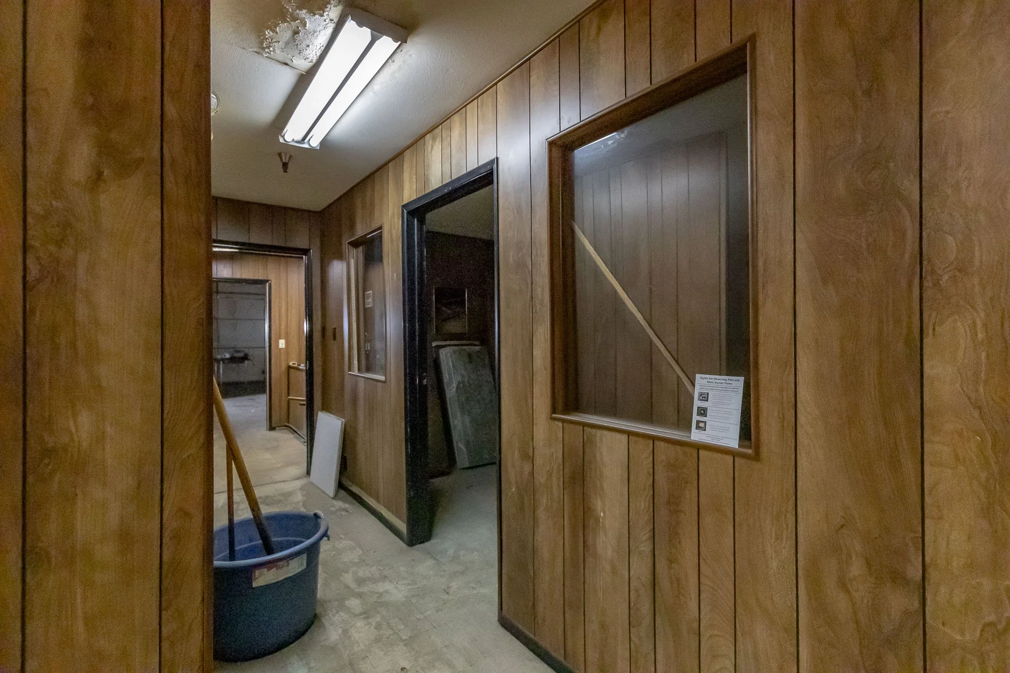 A hallway with wood-paneled walls, fluorescent ceiling light, and a mop with a bucket on the floor. There is a black door and a window with a sign inside a wooden frame on the right wall.