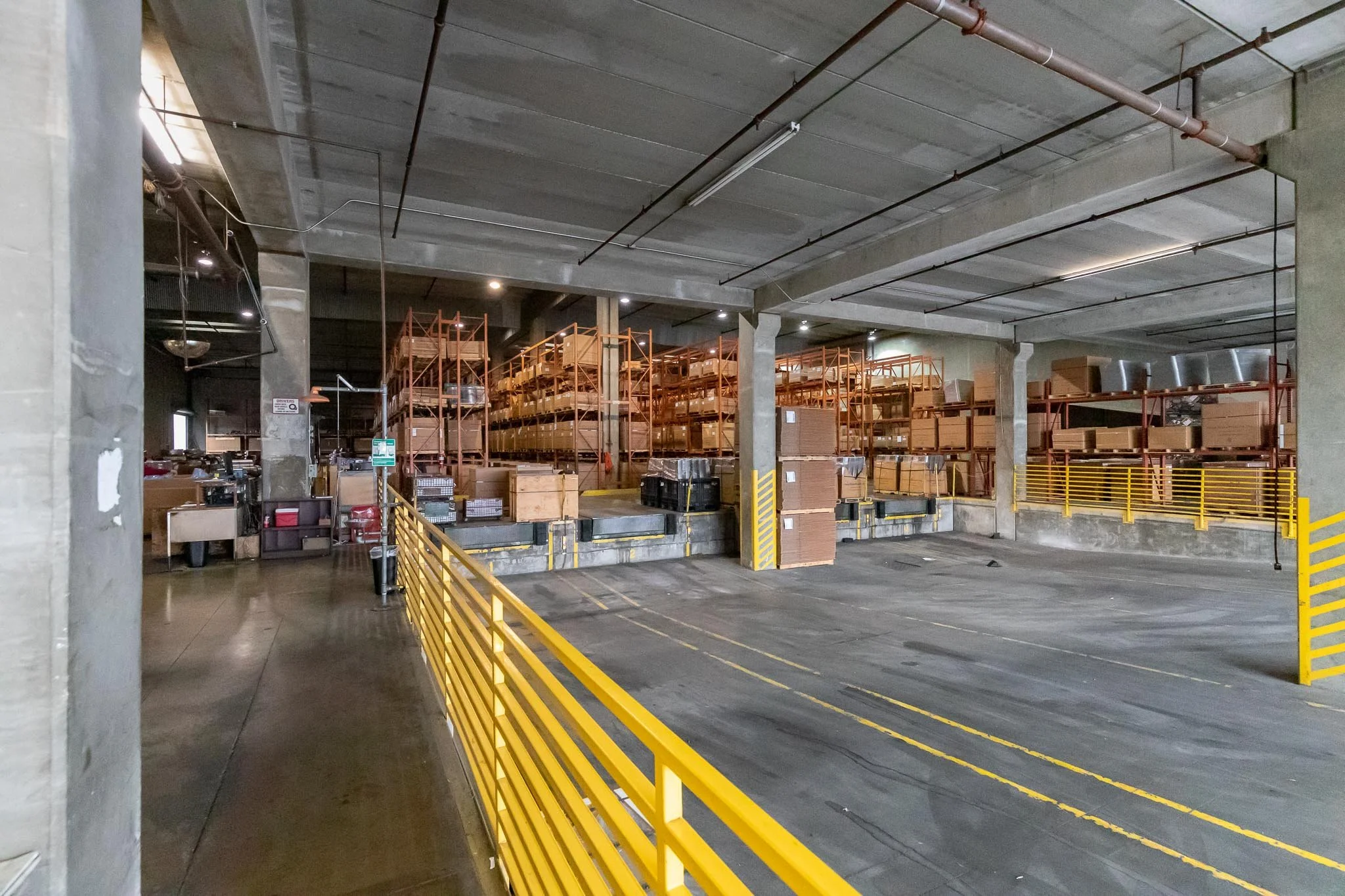 Empty warehouse loading dock with yellow safety rails, stacked cardboard boxes on orange shelving units, and an industrial ceiling with overhead lighting.