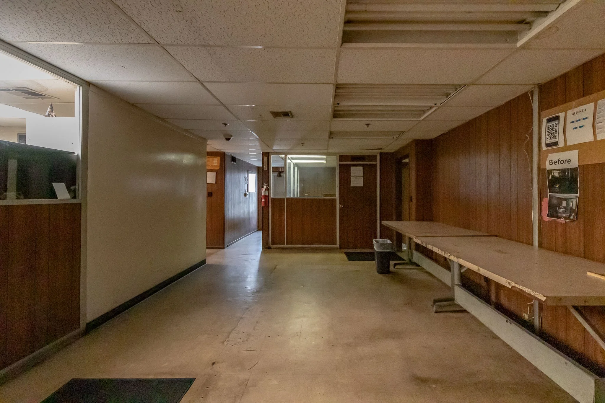 Empty hallway with wood-paneled walls, a table with a trash bin underneath, and a sign on the wall indicating a before renovation state.