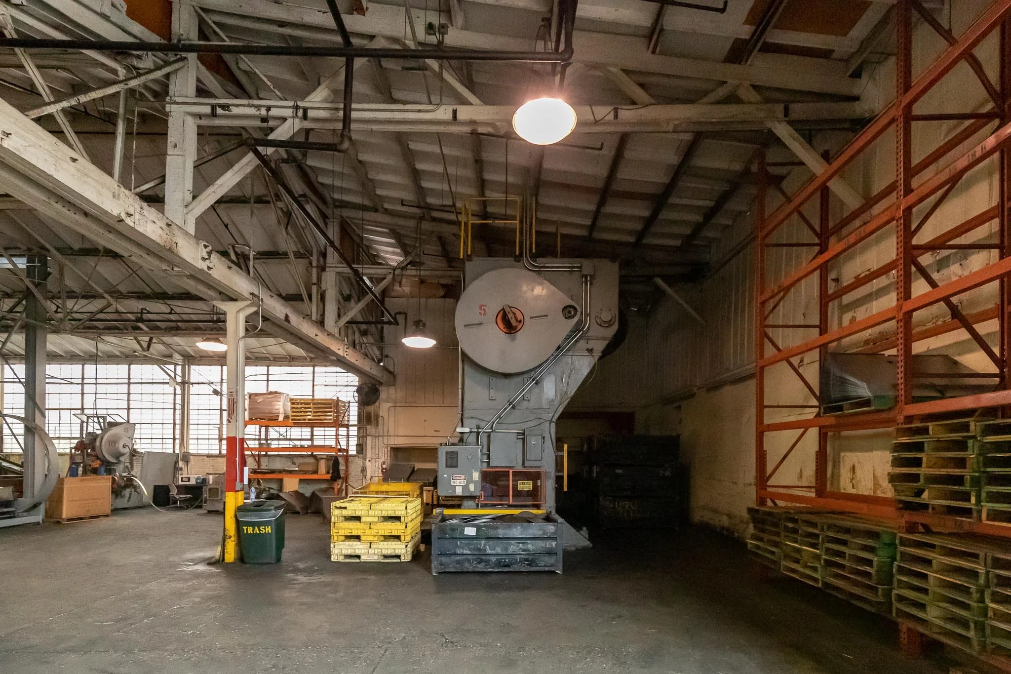 Industrial warehouse with large machinery, wooden pallets stacked on orange shelves, and a trash bin labeled 'TRASH' in the foreground.