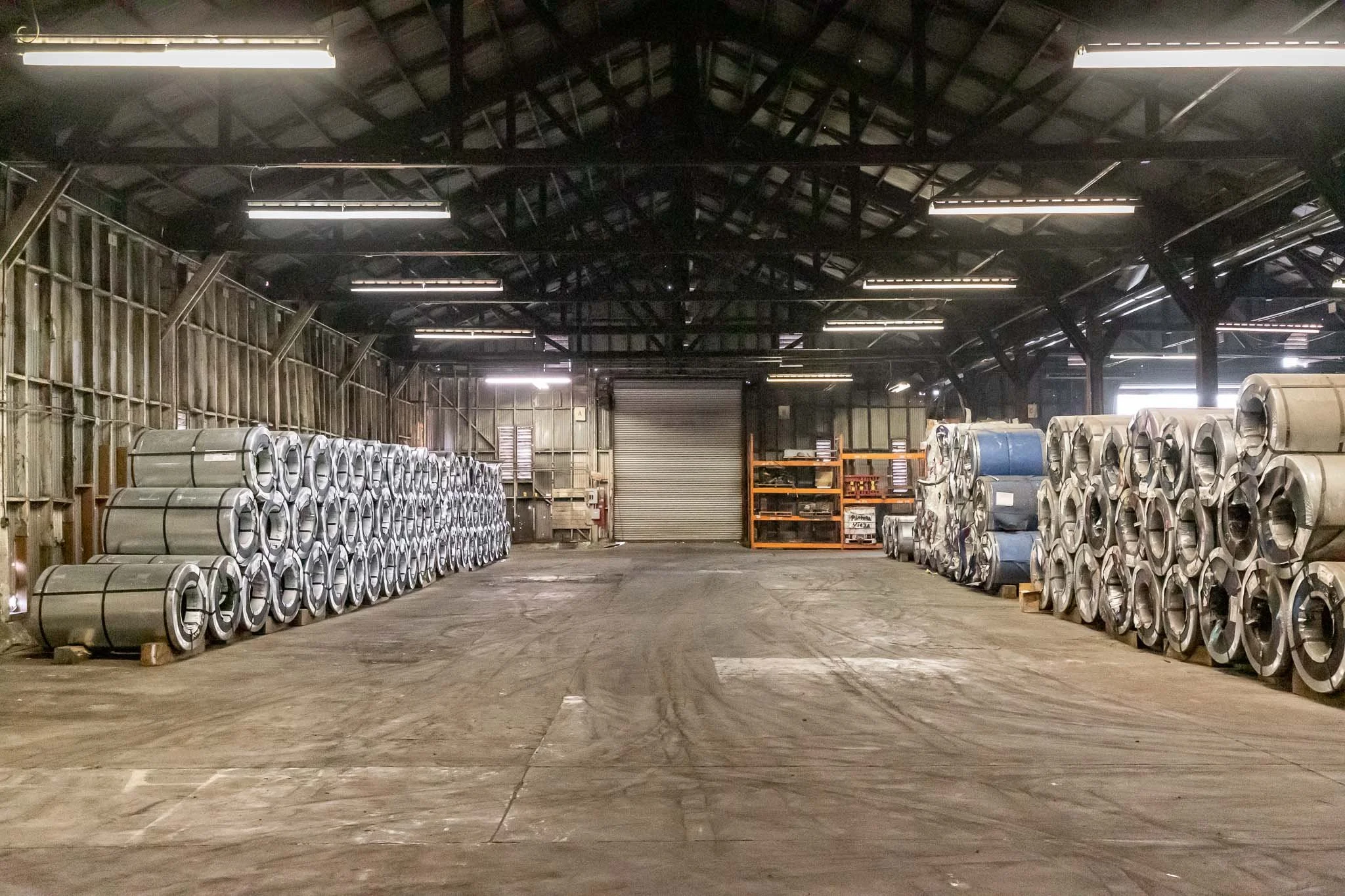 Interior of a warehouse with metal coils stacked on both sides, a roller shutter door at the back, and industrial shelving units on the right side.