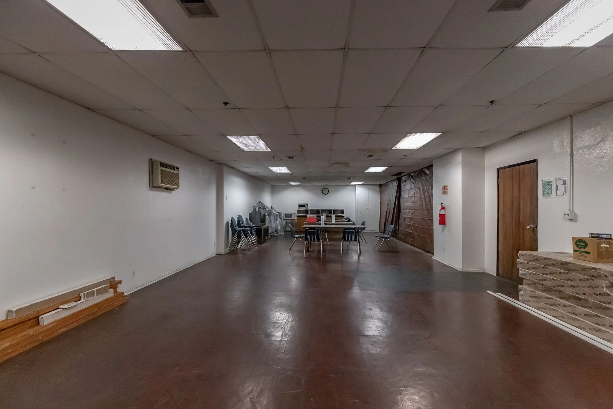 Empty room with a wooden floor, white walls, ceiling lights, and some chairs and tables in the back. There's a brick structure on the right side and a few stacked chairs near the back wall.