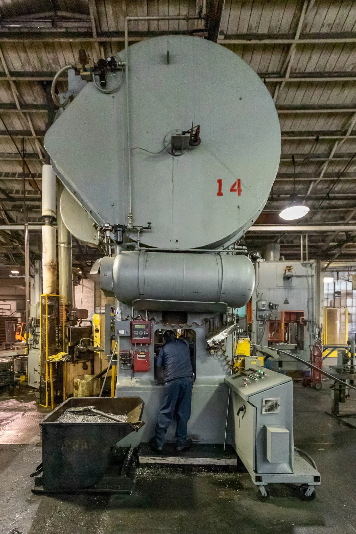 A worker in a factory operating a large industrial machine, with a control panel and surrounding machinery, inside a spacious industrial building.