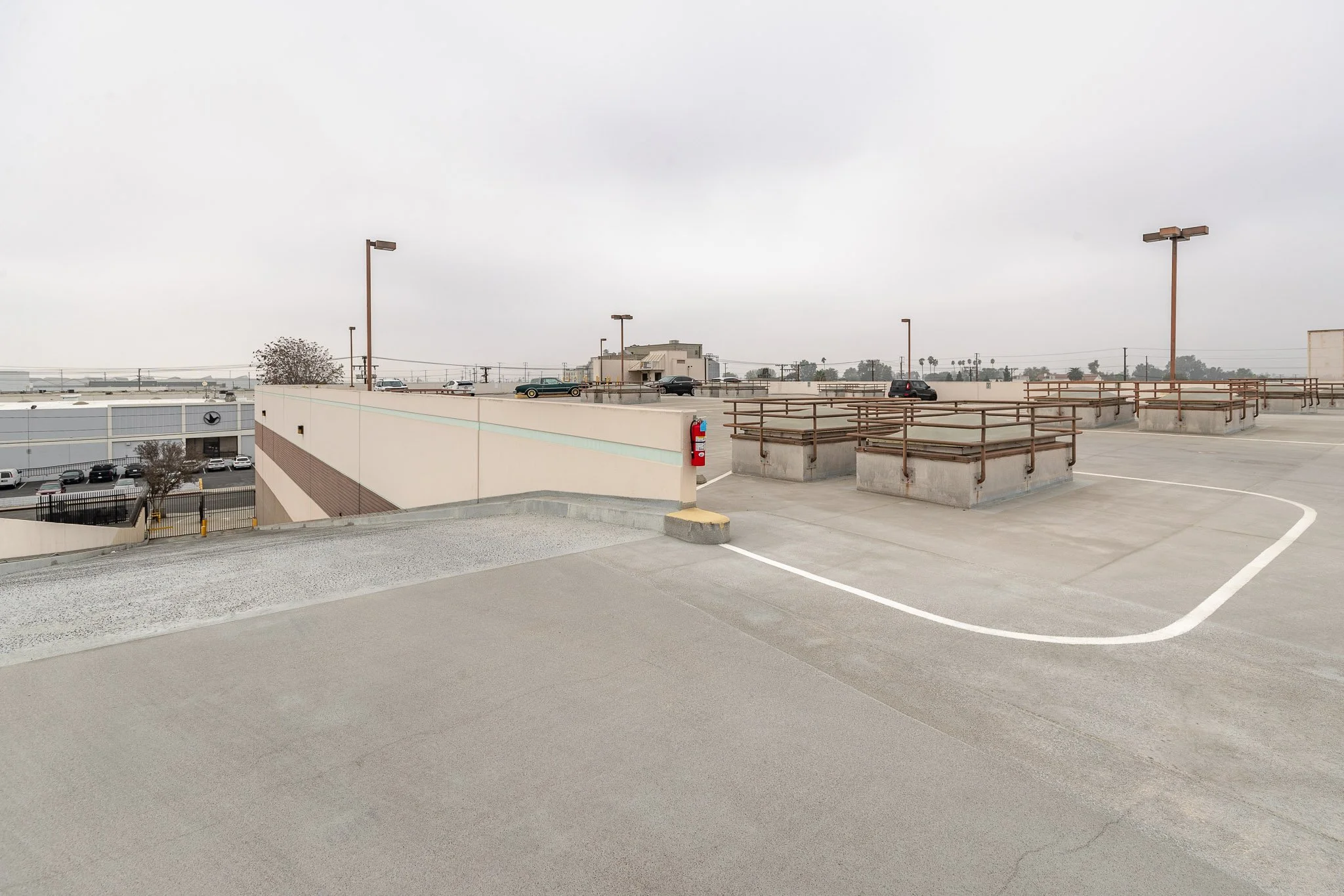 Empty rooftop parking lot with a few cars, safety railings, and a fire extinguisher on the wall, overcast sky.