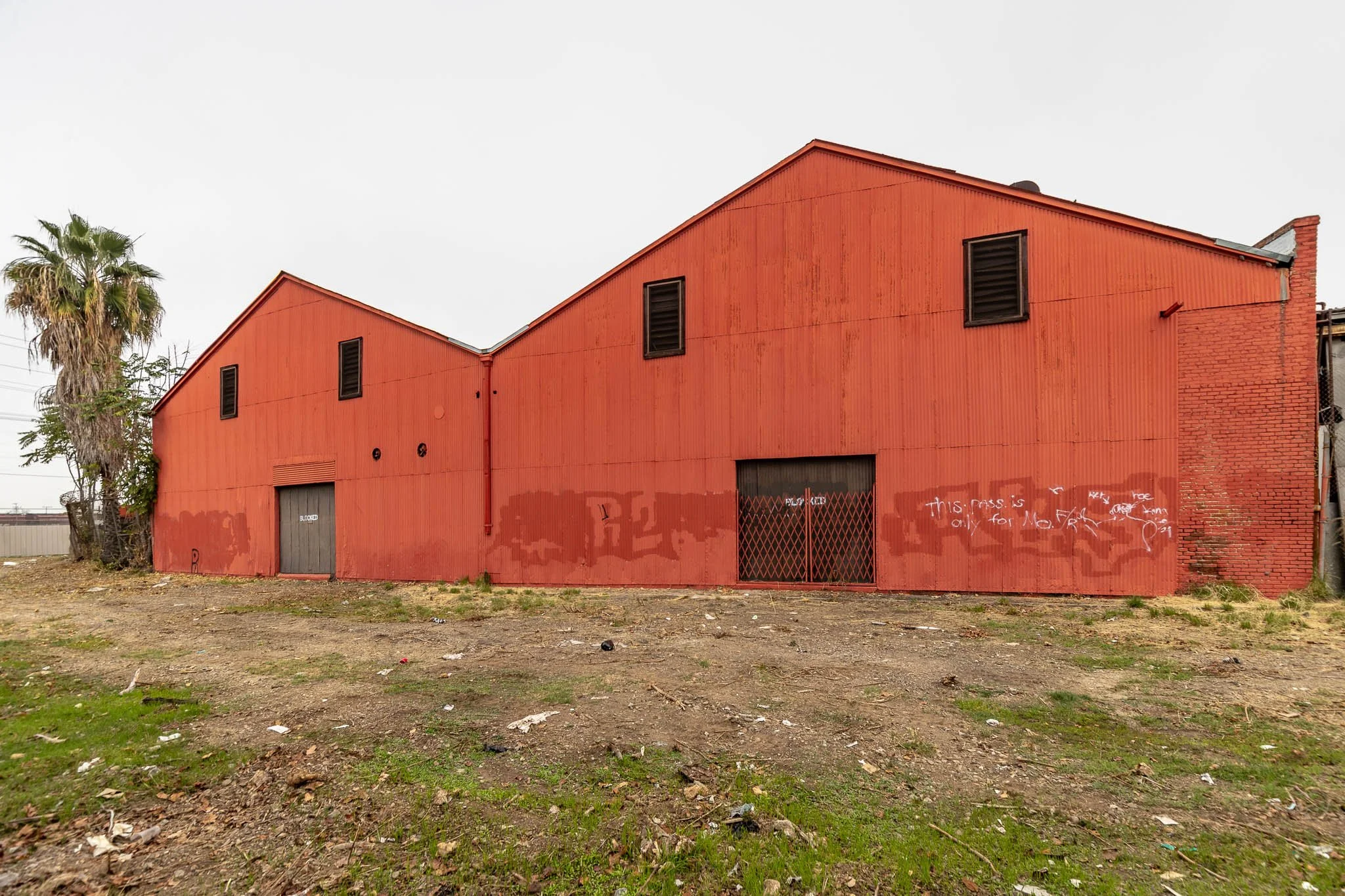 An old red warehouse with graffitied walls, black vents, metal doors, and surrounding dirt and grass.