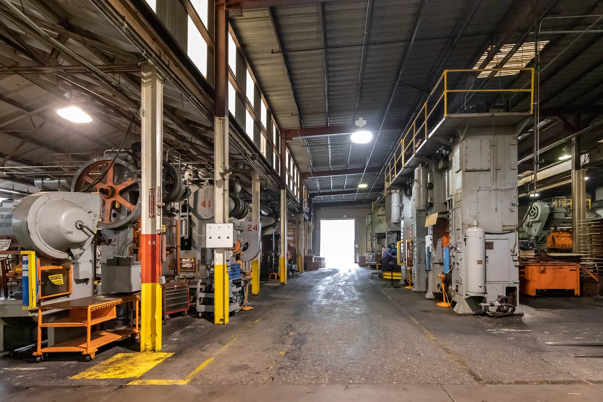 Industrial workshop with large machinery, tools, and a bright open door at the end of the space.