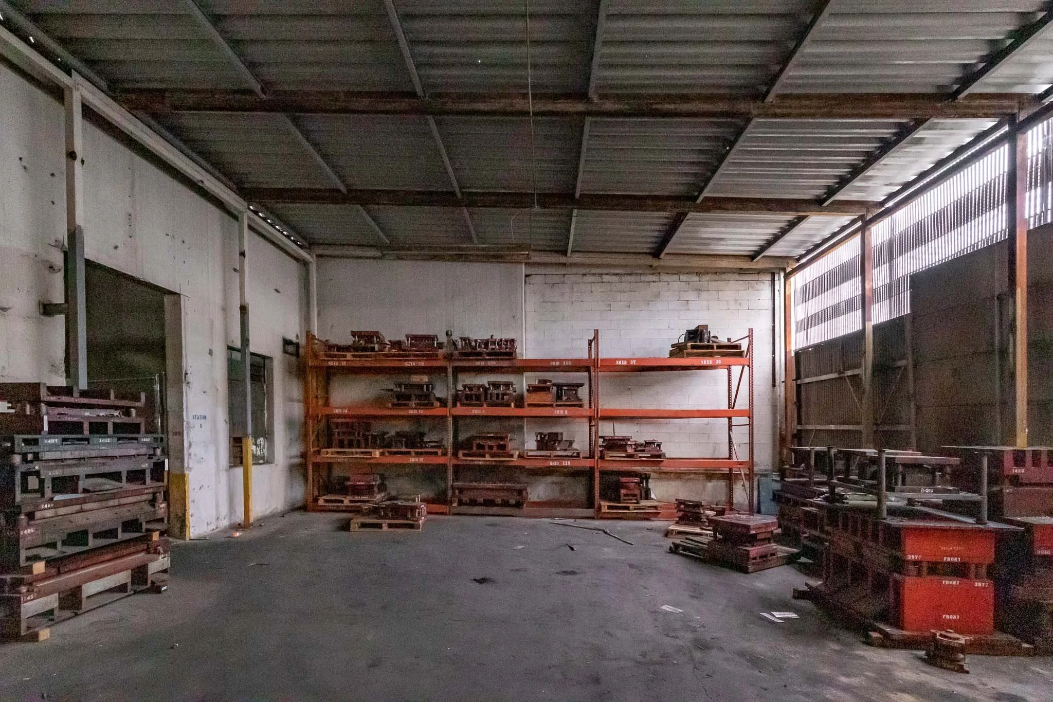 Interior of a warehouse with metal shelving units and stacked wooden pallets, some containing metal or wooden objects, against white walls and a corrugated metal roof.