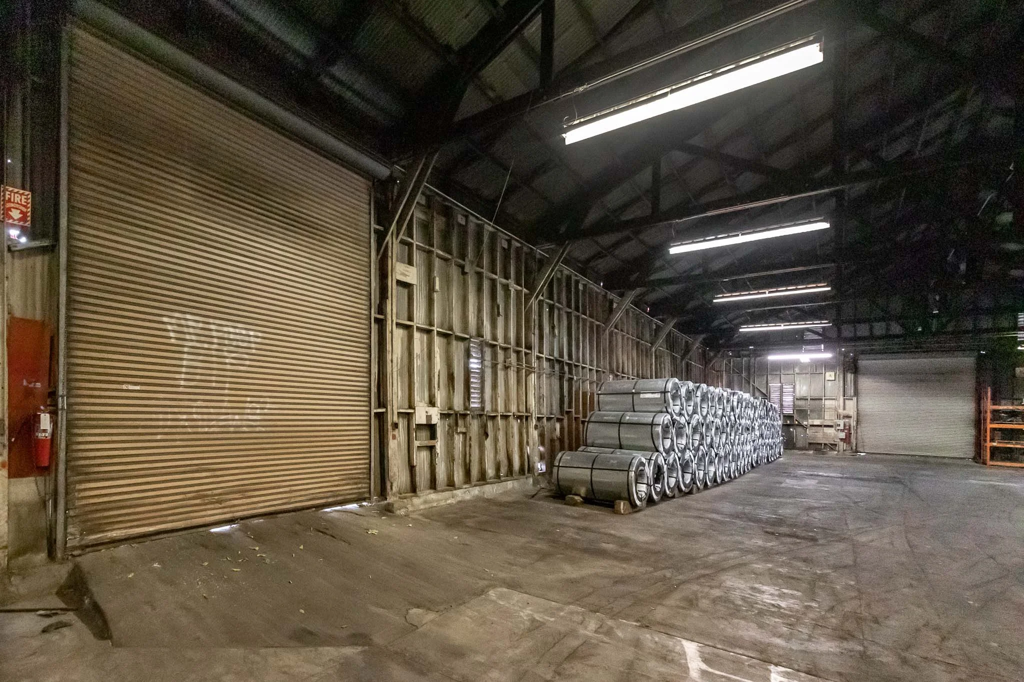 Interior of a warehouse with stacked metal rolls on the right side and a closed rolling shutter door on the left.