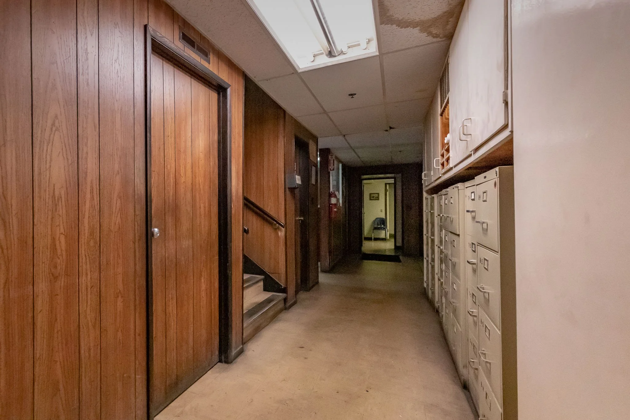 A hallway with wooden panel walls, lockers on the right, a door on the left, and a chair visible through an open doorway at the end.