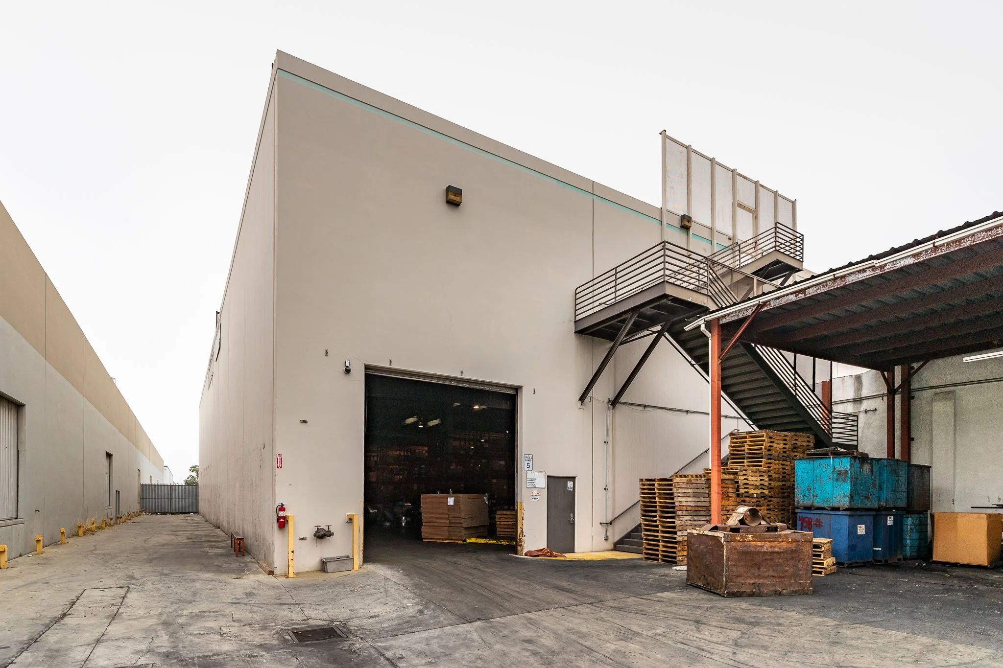 An industrial building with a loading dock, metal stairs, stacked pallets, and storage containers outside.