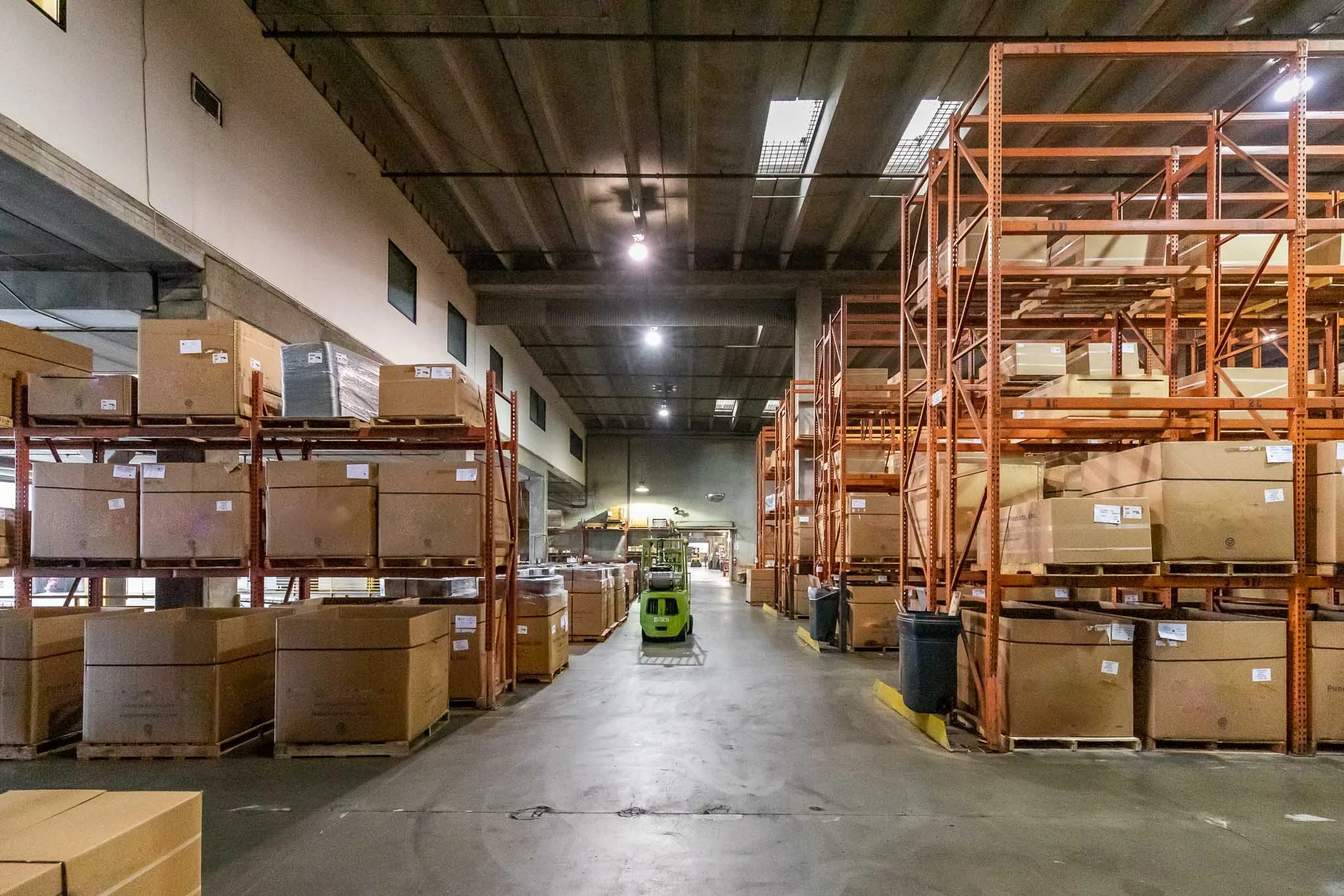 Warehouse with orange metal shelving filled with large cardboard boxes, a green forklift in the center, and industrial lighting overhead.