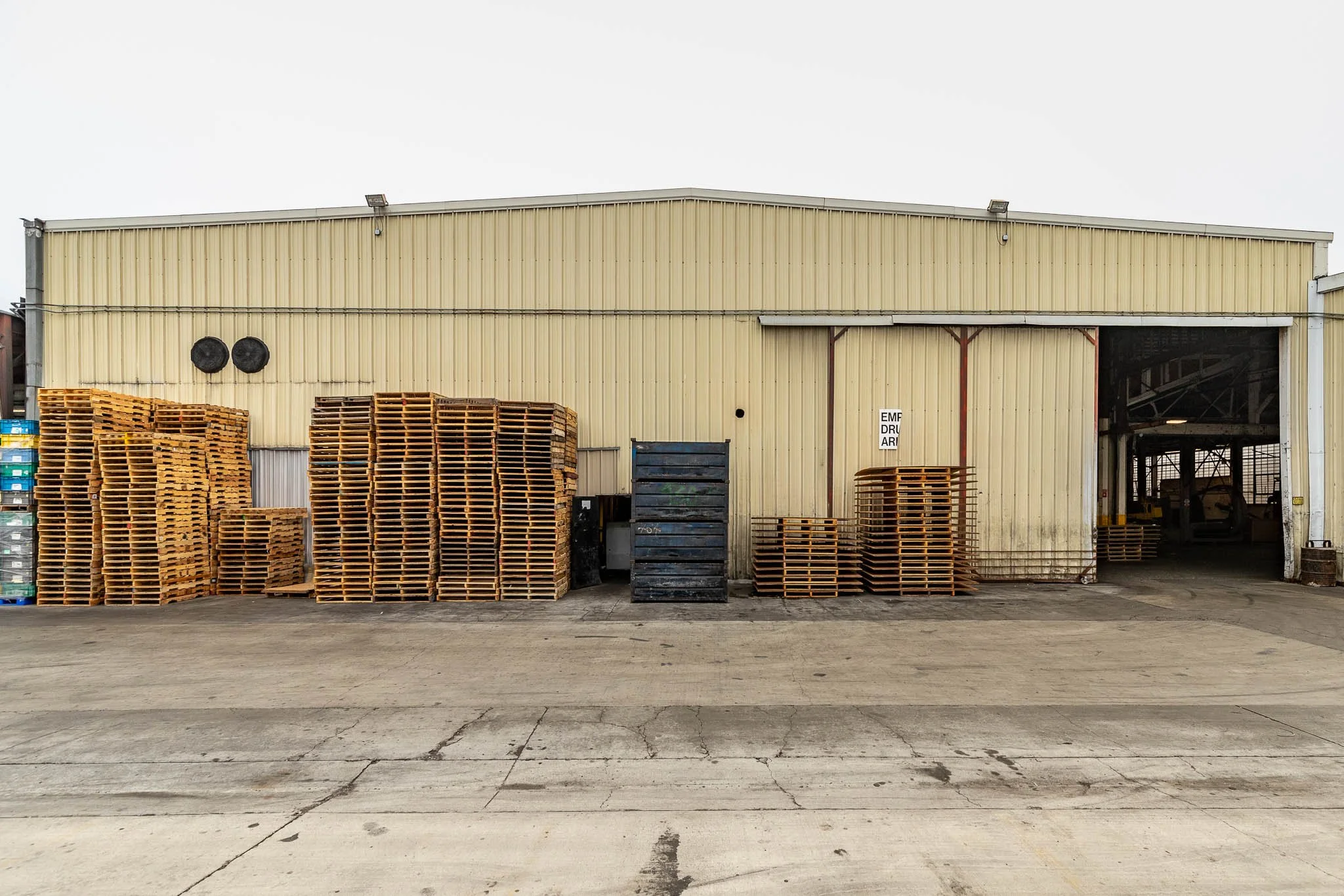 Stacks of wooden pallets outside a warehouse with a yellow metal exterior and an open sliding door.