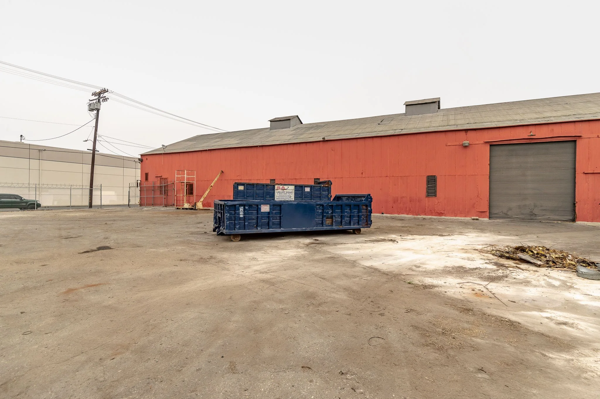 Empty lot with a large blue dumpster in front of a red warehouse building and a closed rolling door, with some debris on the ground to the right.