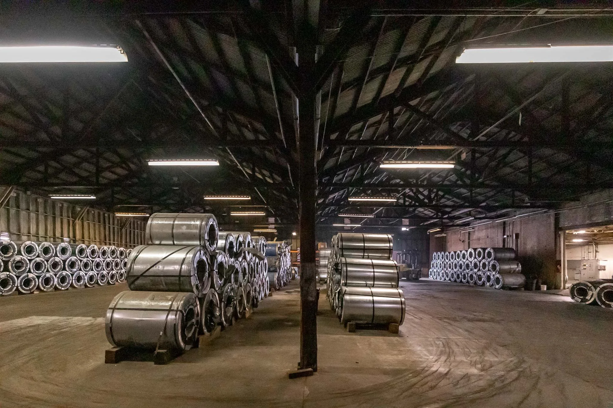 Steel coils stored inside an industrial warehouse with a high ceiling and fluorescent lighting.