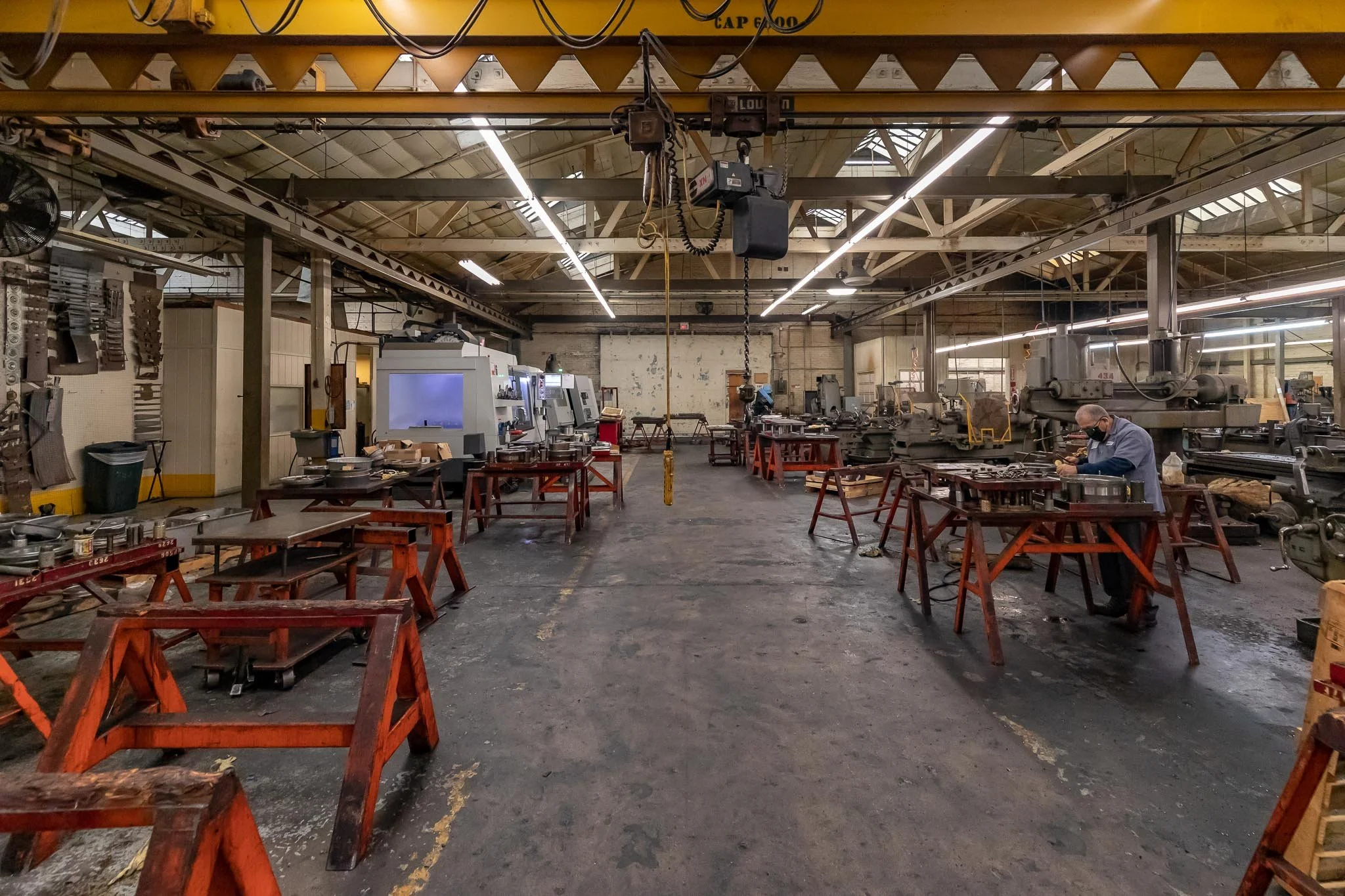 An industrial workshop with machinery, workbenches, and a worker wearing a mask and protective gear, engaged in metalworking or machining work.