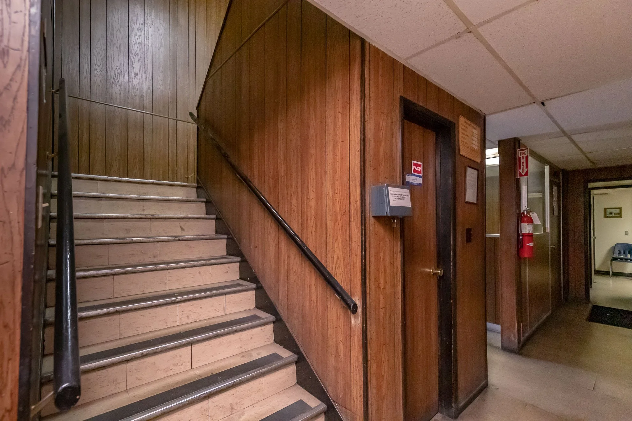 Interior view of a stairway with wood paneling walls, an elevator, a fire extinguisher, and a waiting area with chairs.