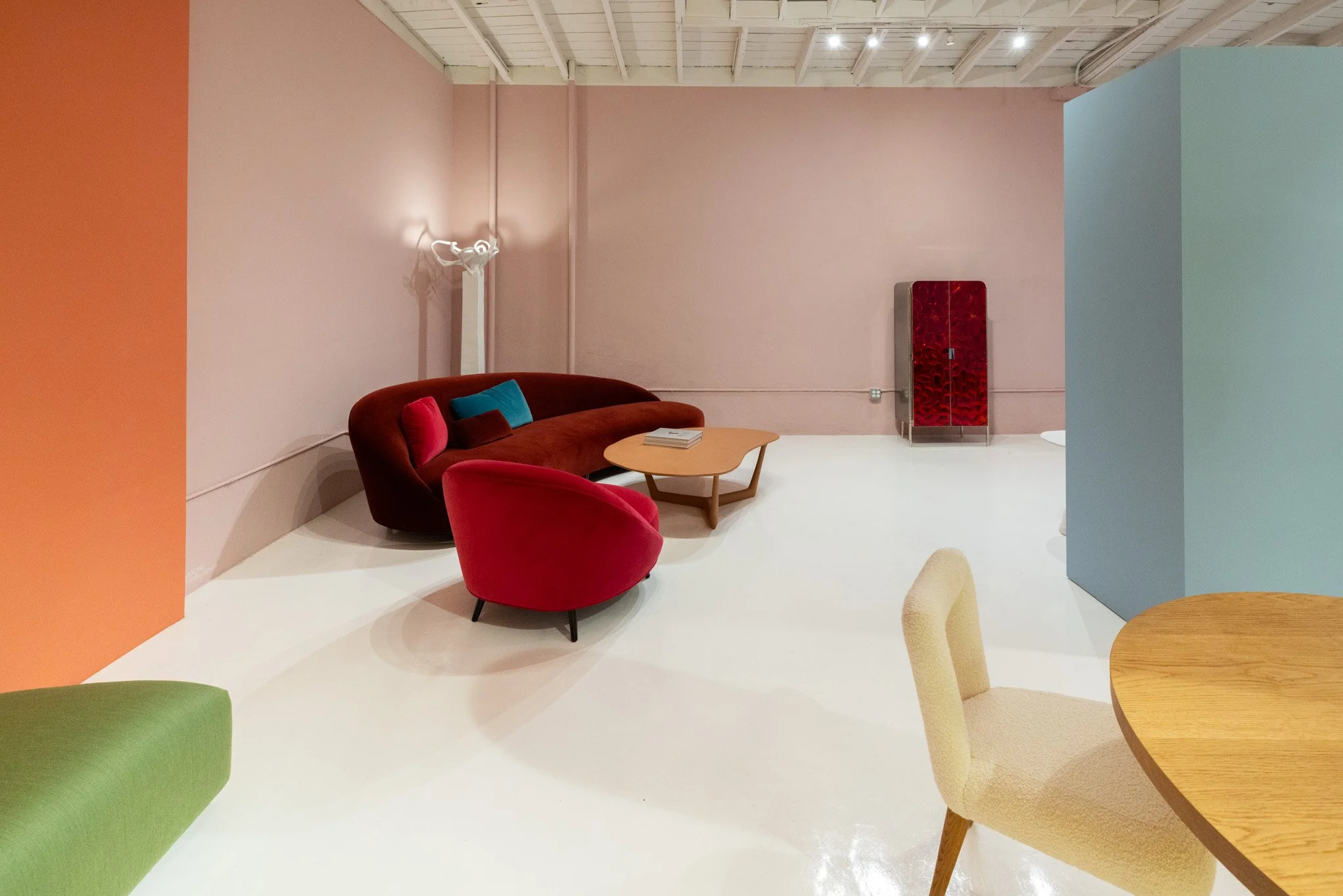 Modern interior room with a pink wall, a red sofa, a red armchair, a small wooden coffee table with books, and a corner red cabinet against the wall. There are also partially visible chairs and a pastel-colored wall.