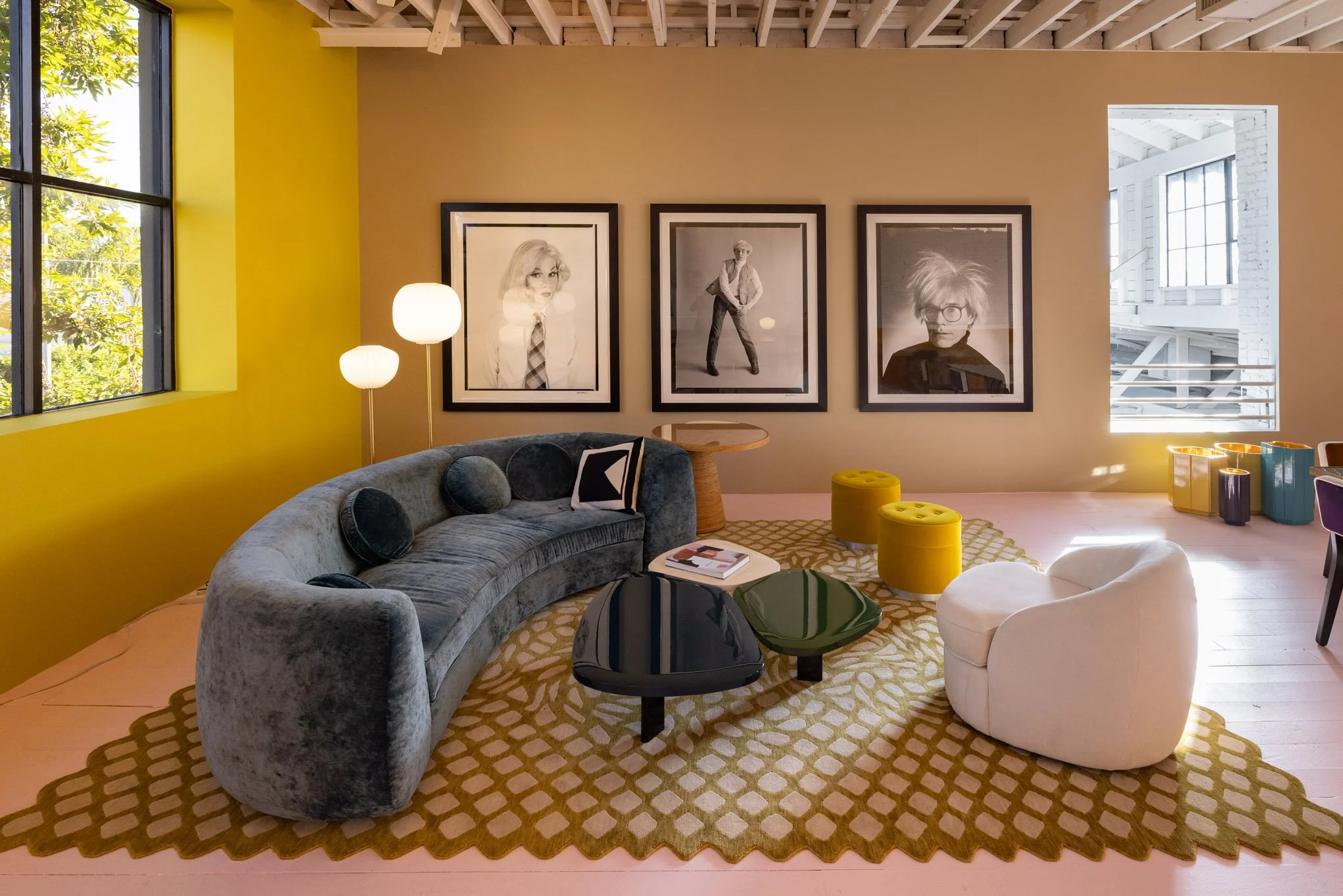 Modern living room with a curved gray velvet sofa, black and yellow ottomans, white armchair, abstract patterned rug, artwork of three black-and-white portraits on the wall, large window with natural light, and a yellow wall.