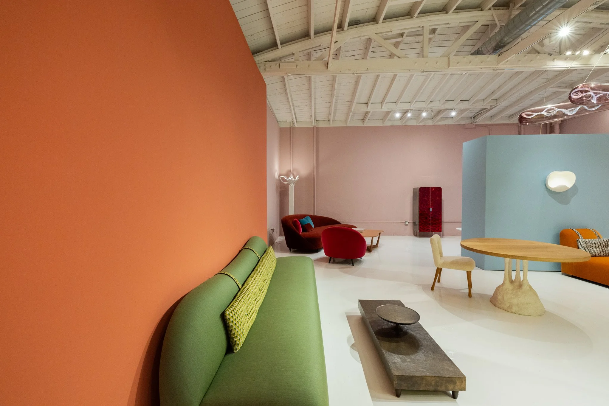Modern interior with colorful seating arrangements, including a green sofa, red and orange chairs, and a unique wooden table with a sculptural base, within a spacious room with high wooden ceilings and pastel-colored walls.