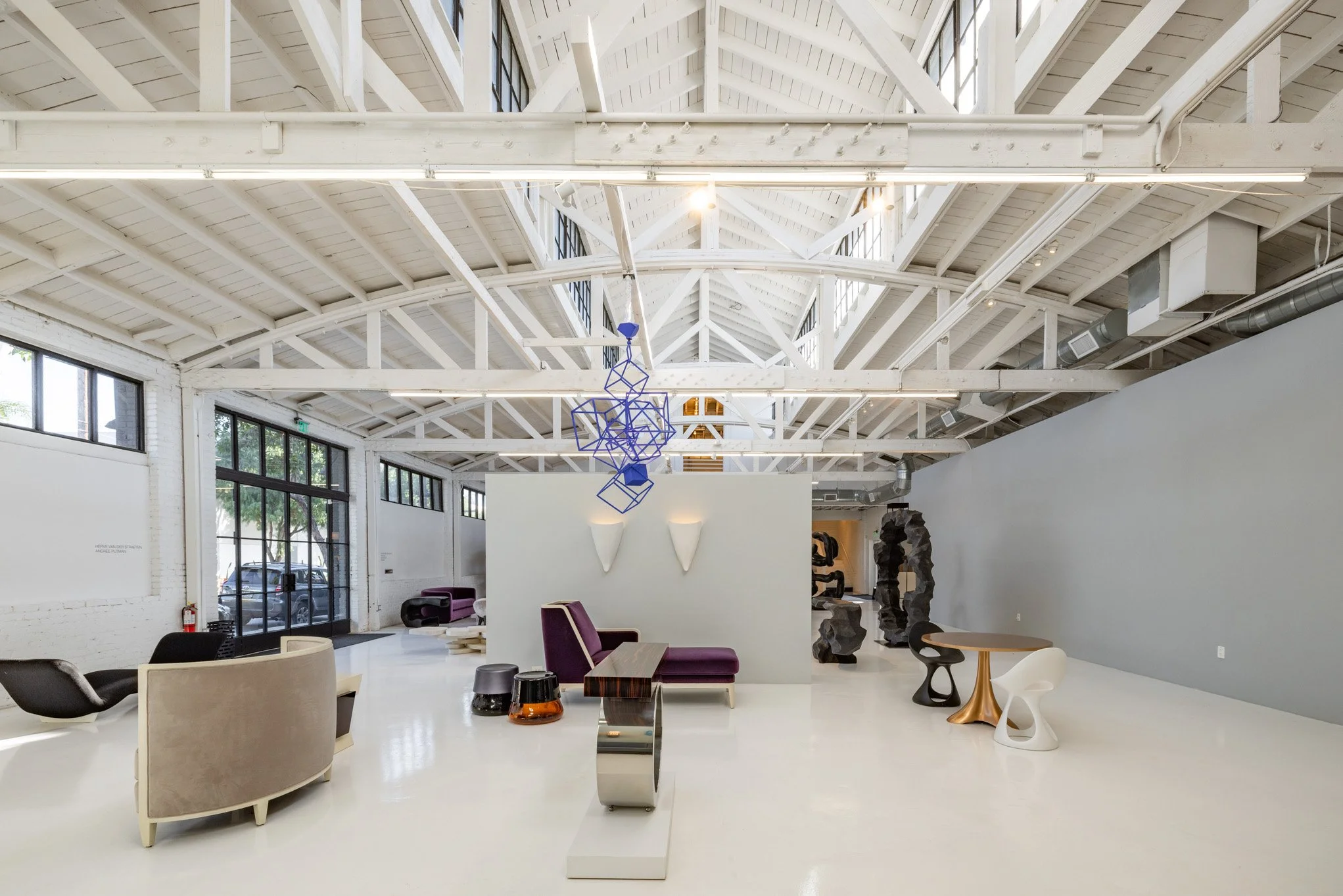 Interior of a modern gallery with white walls, high vaulted ceiling with exposed beams, and contemporary furniture including chairs, tables, and sculptural art pieces.