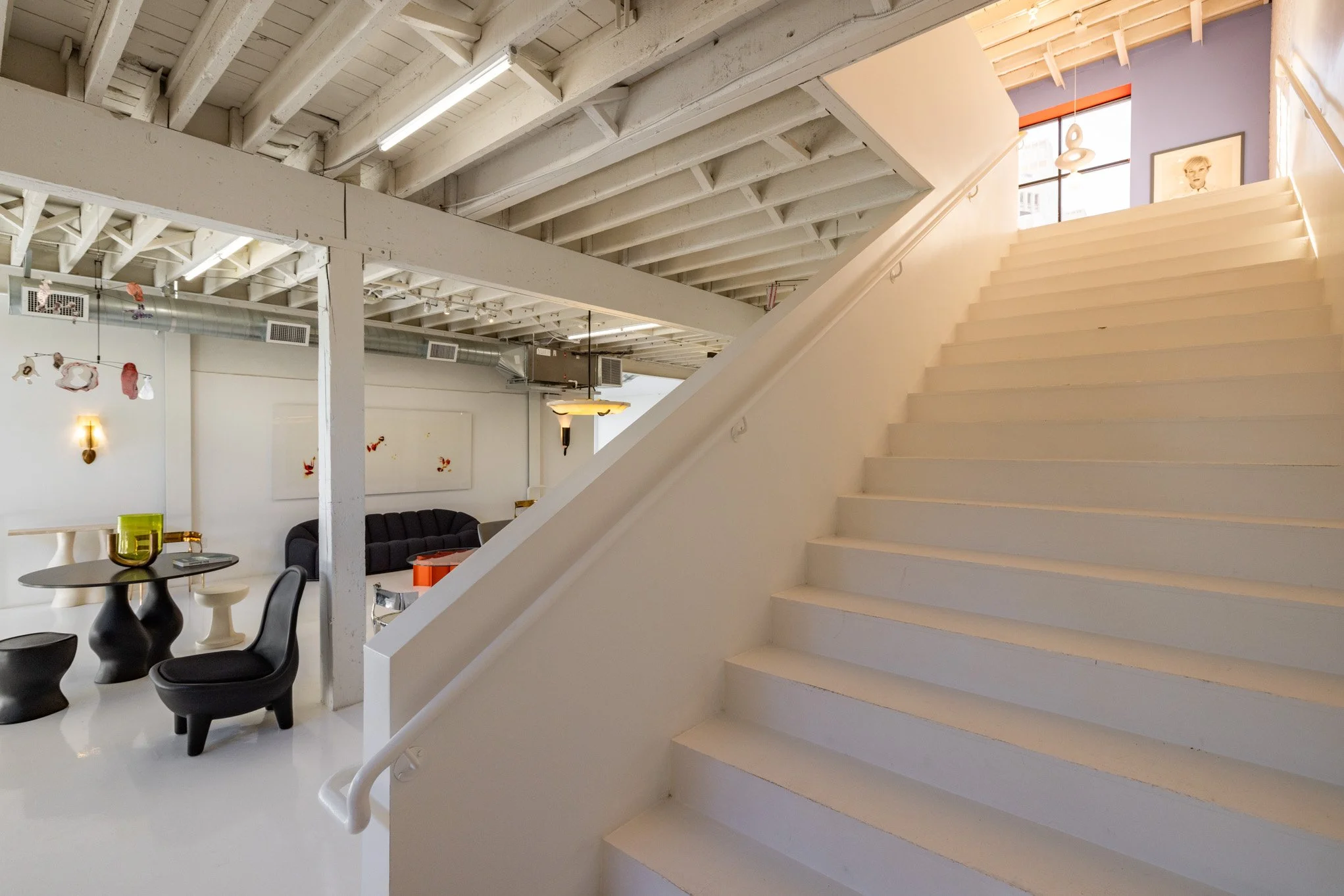 Interior of a modern, industrial-style space with white stairs, exposed beams, and artwork, featuring seating areas and high ceilings.
