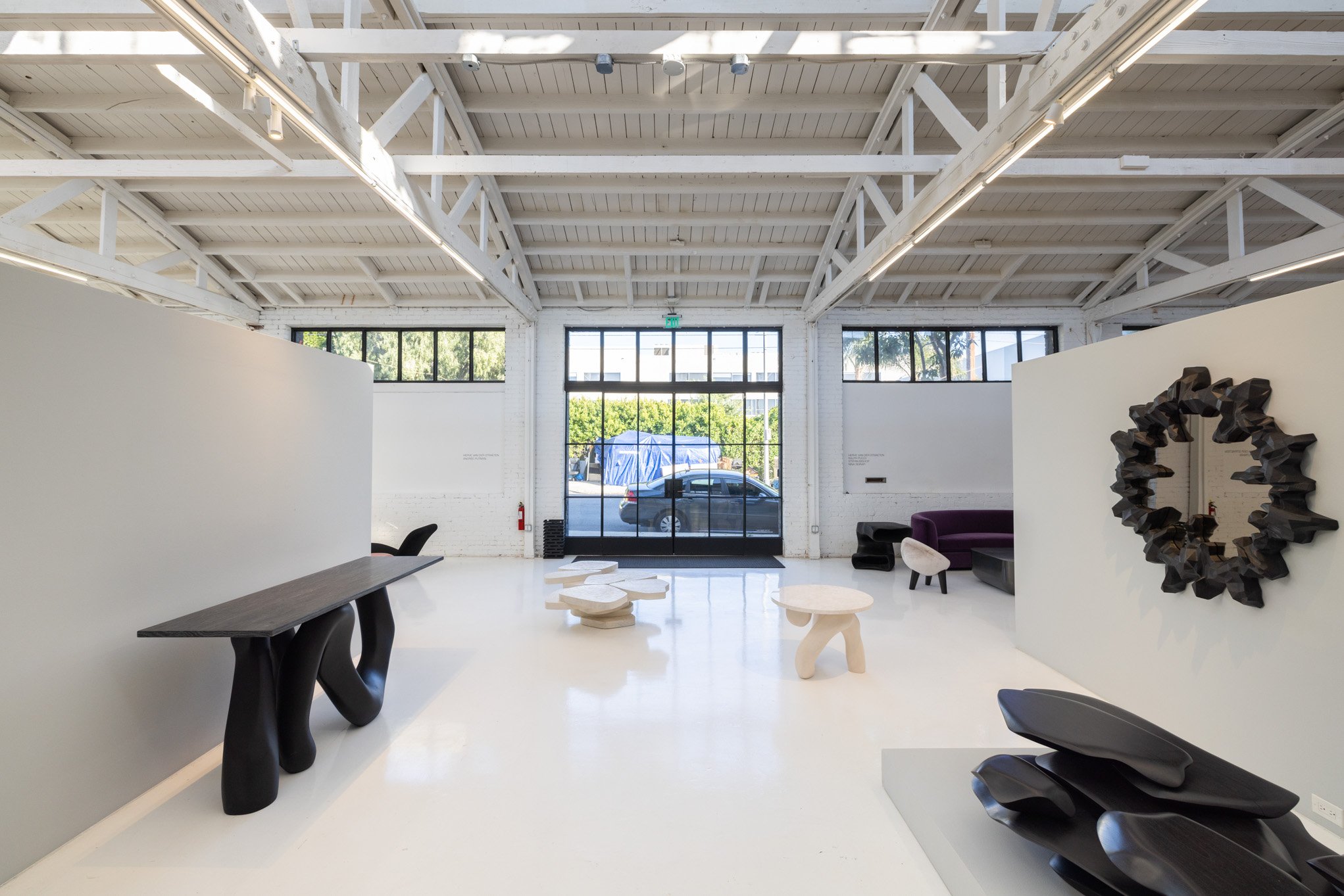 A modern gallery space with white walls, high ceilings with exposed white beams, large front windows, and contemporary furniture including a black sculpted bench, a purple sofa, white and black decorative tables, and a decorative black circular mirro