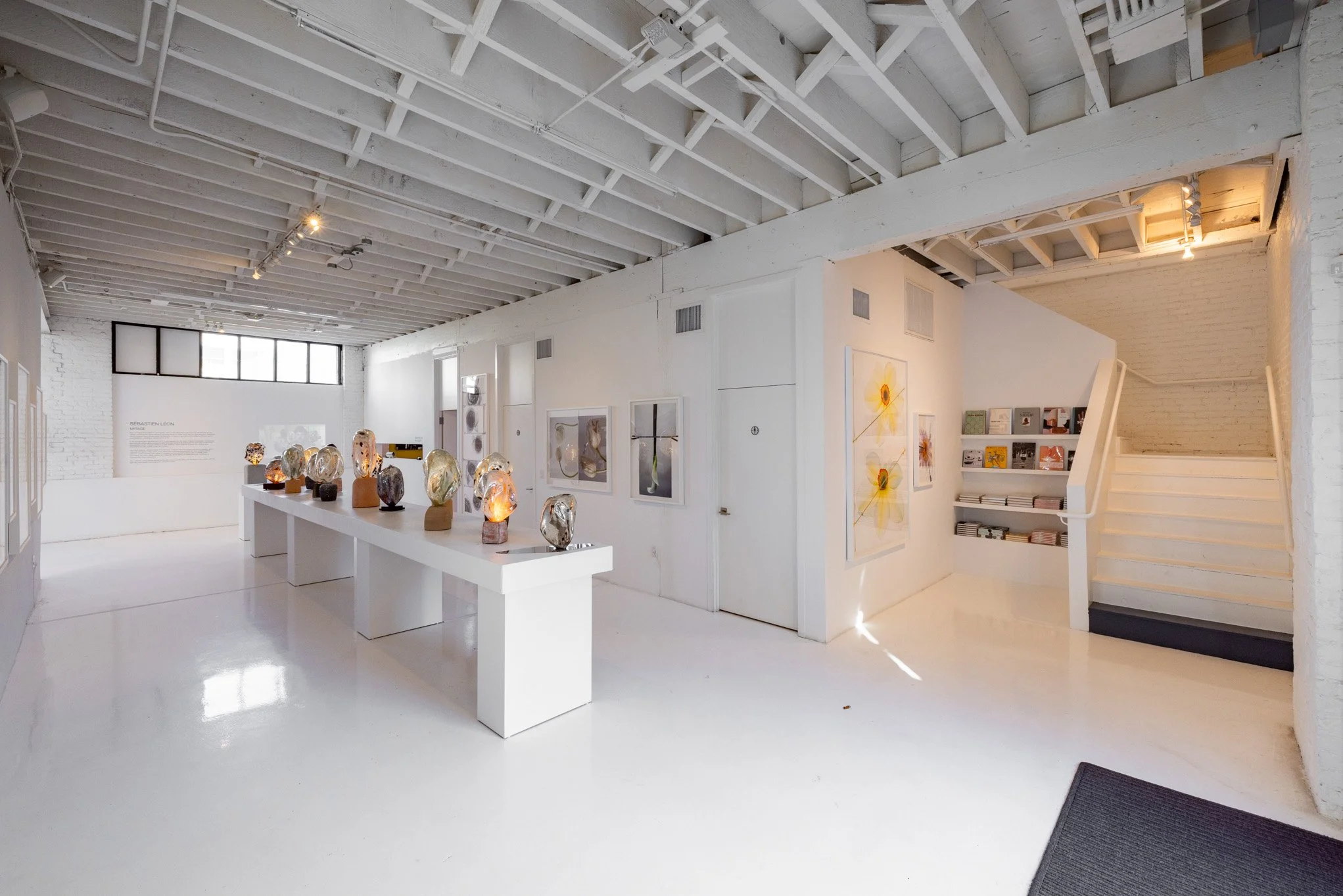 Modern art gallery with white walls and floors, displaying sculptures on white tables and framed artwork on the walls, with a staircase on the right side.