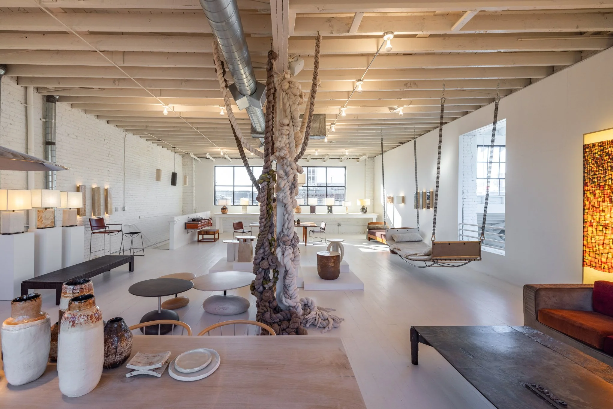 An art gallery with white brick walls and wooden ceilings, featuring sculptures, pottery, and a hanging rope art installation.