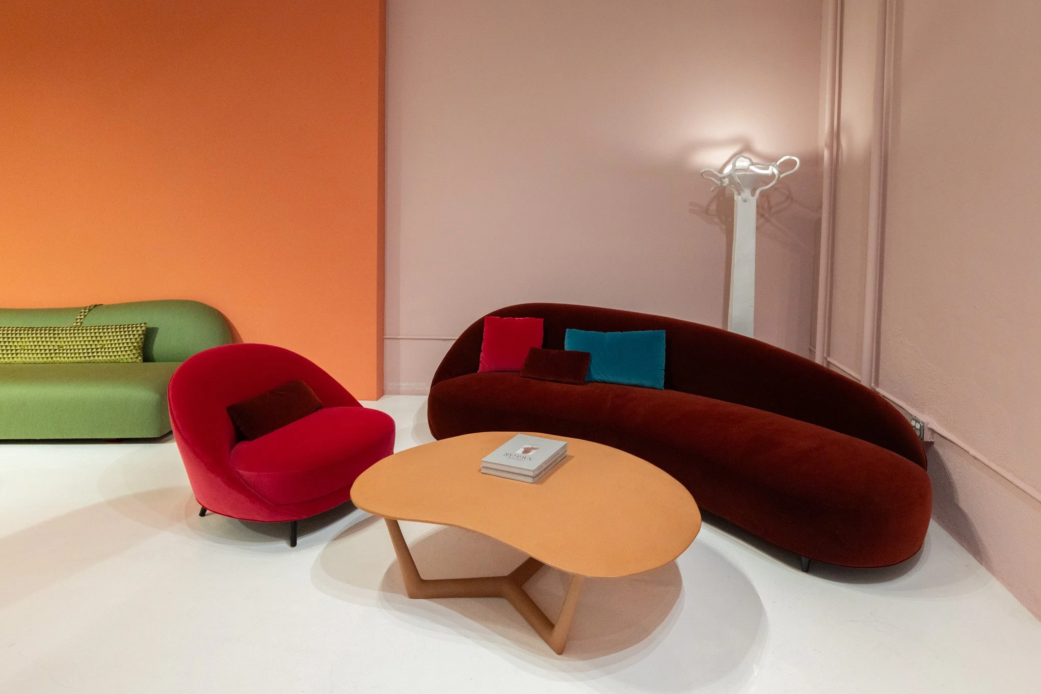 Colorful modern furniture including a red armchair, a green sofa, a brown curved sofa with pillows, and a light wood coffee table in a contemporary room with a peach and white wall and a coat rack.