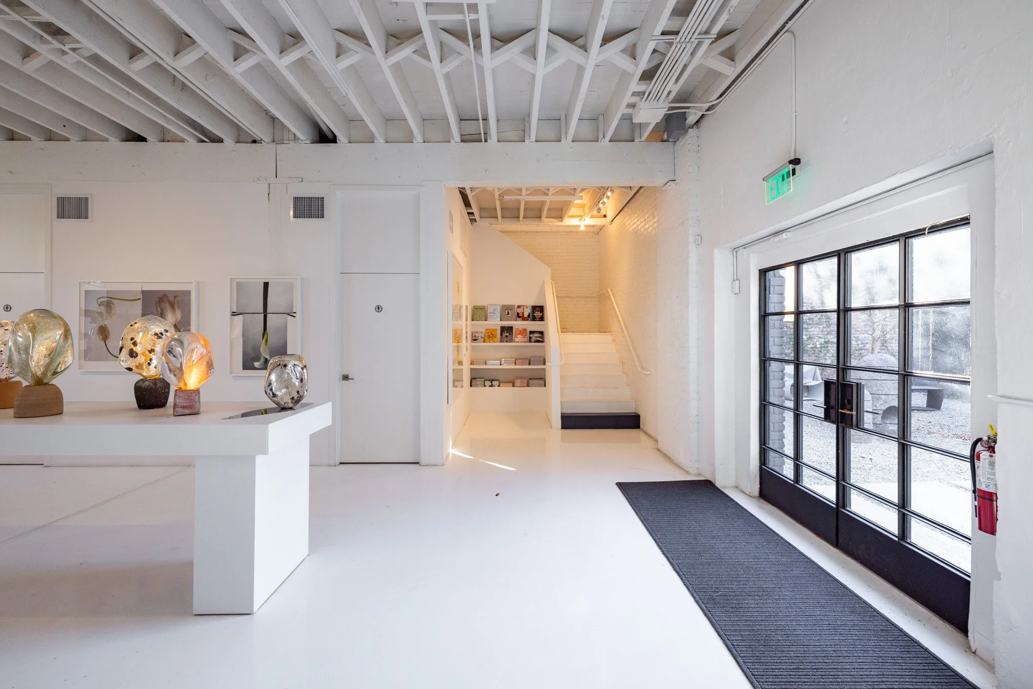 Interior of a modern art gallery with white walls and ceiling, display table with glass art sculptures, staircase with bookshelves, glass storefront door with outdoor seating visible through the glass.