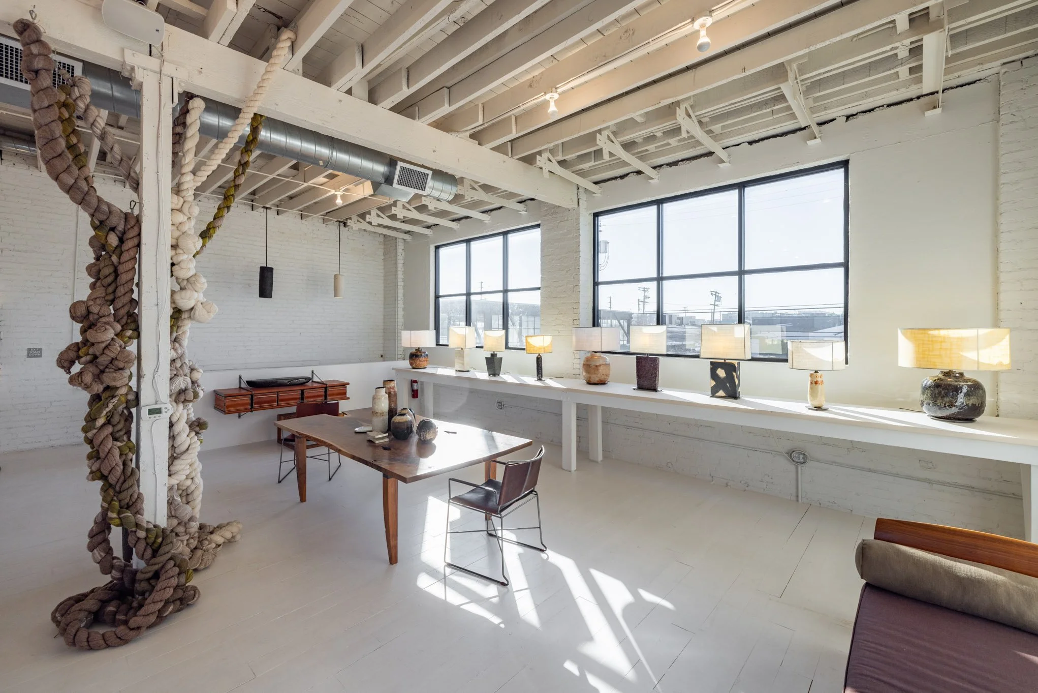 An art gallery with white brick walls, large windows, and art pieces including lamps and sculpture. Sunlight streams through the windows, illuminating the space.