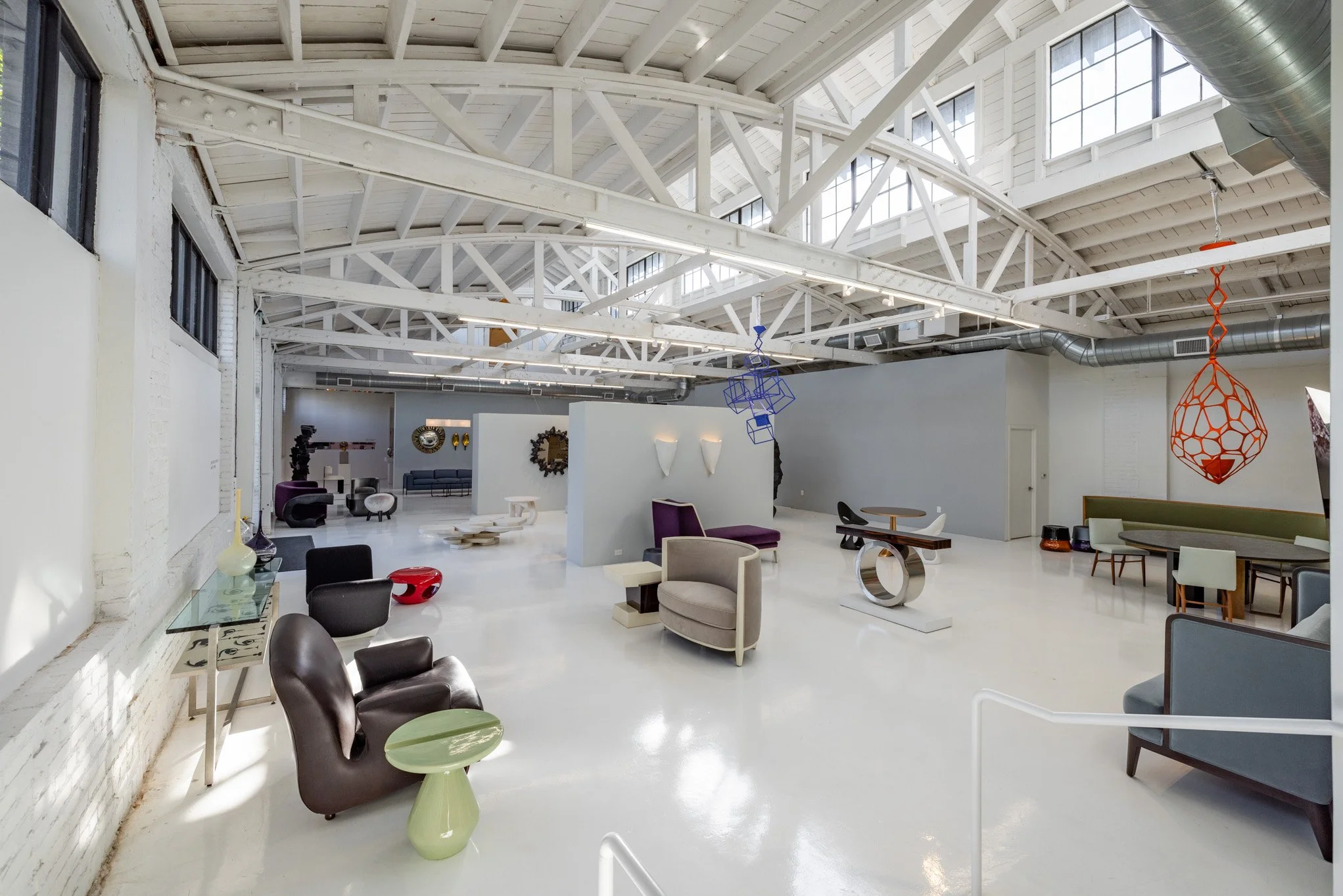 A spacious, modern art gallery with white painted walls, high vaulted ceilings with exposed beams, and a glossy white floor. The gallery features various contemporary furniture and artistic sculptures, including colorful wireframe hanging art pieces 