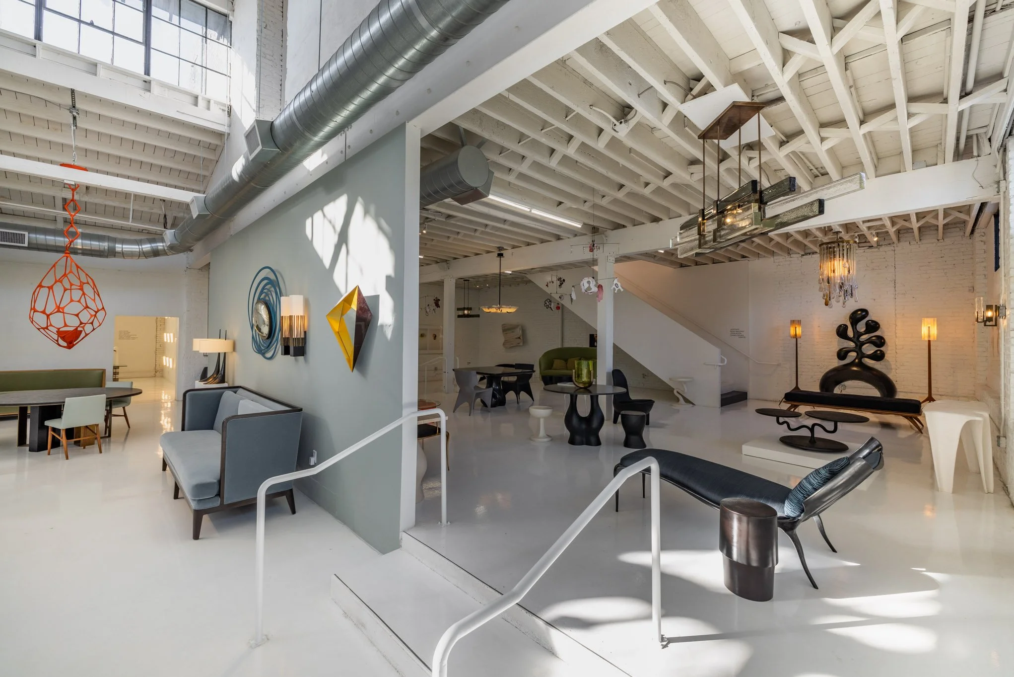 Interior of an art gallery or showroom with modern furniture, sculptures, and decorative lighting, featuring a white ceiling with exposed beams and ductwork, and a white brick wall.