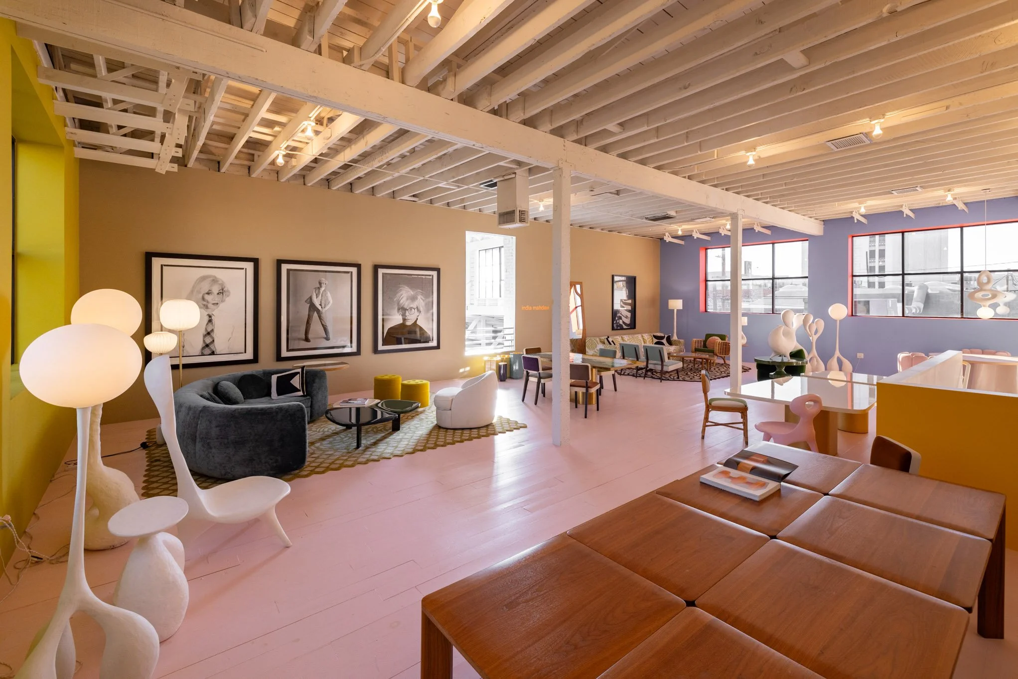 Colorful modern living room with various furniture pieces including couches, chairs, tables, and decorative lighting, featuring large windows and framed artwork on the walls.
