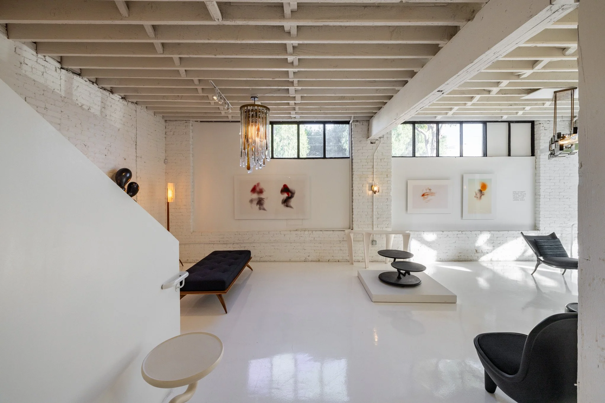 An art gallery with white brick walls, exposed ceiling beams, large windows, modern black and white furniture, and abstract artwork on the walls.