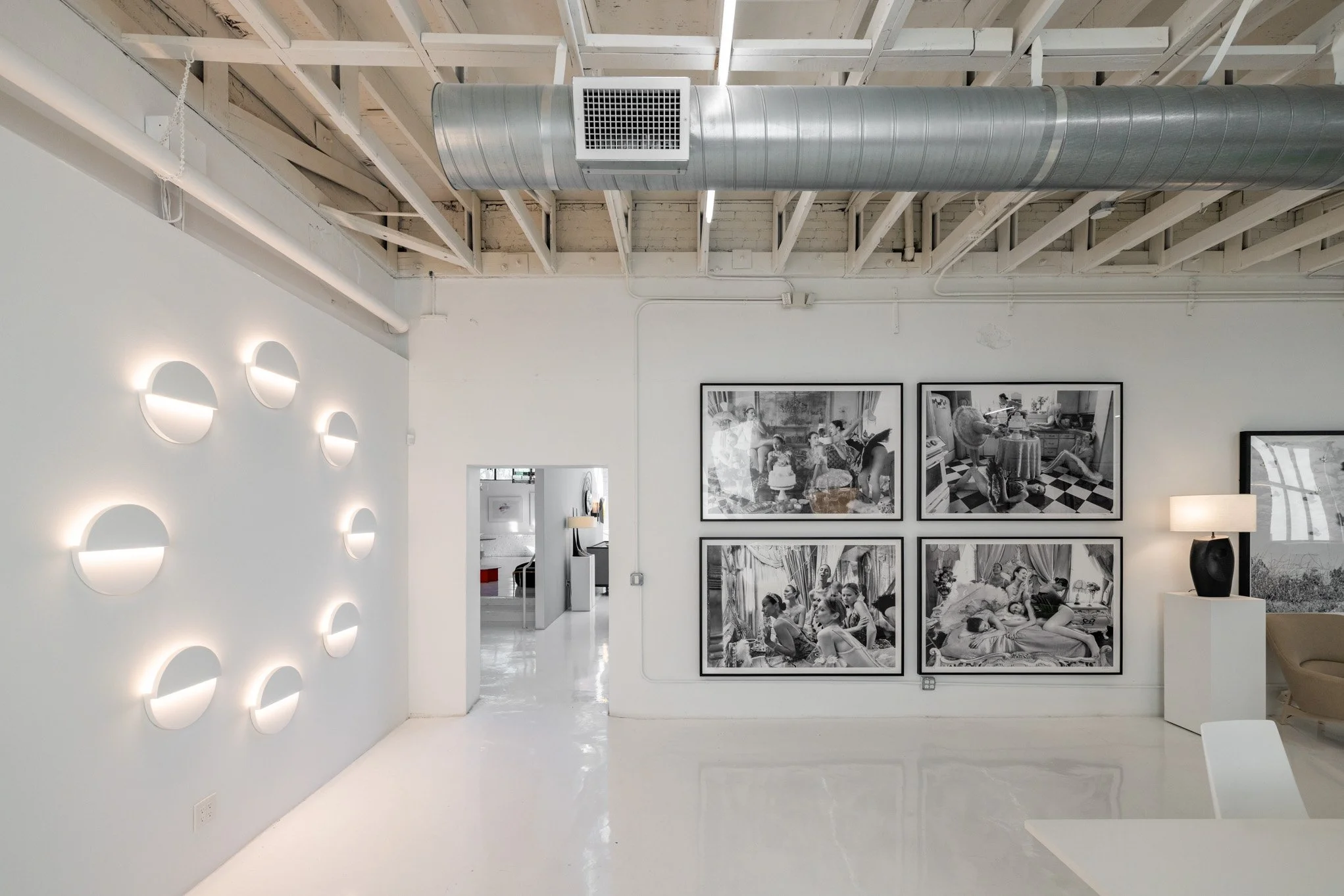 Gallery with black-and-white photographs on the wall, modern lighting on the white wall, exposed ceiling pipes, and minimal furnishings.