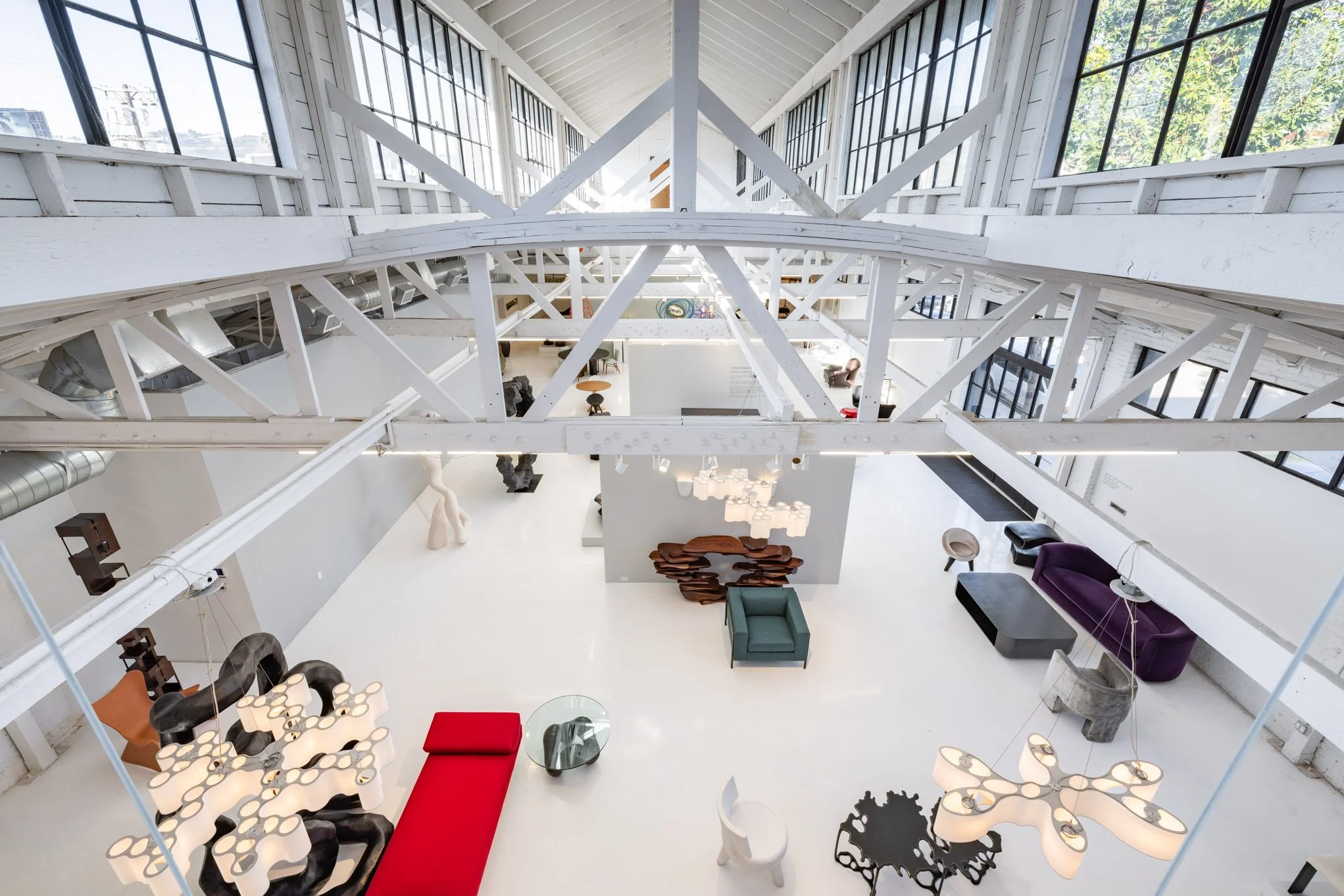 Interior of a modern, industrial-style building with white exposed beams, large windows, and contemporary furniture including colorful sofas, chairs, and stylish light fixtures.