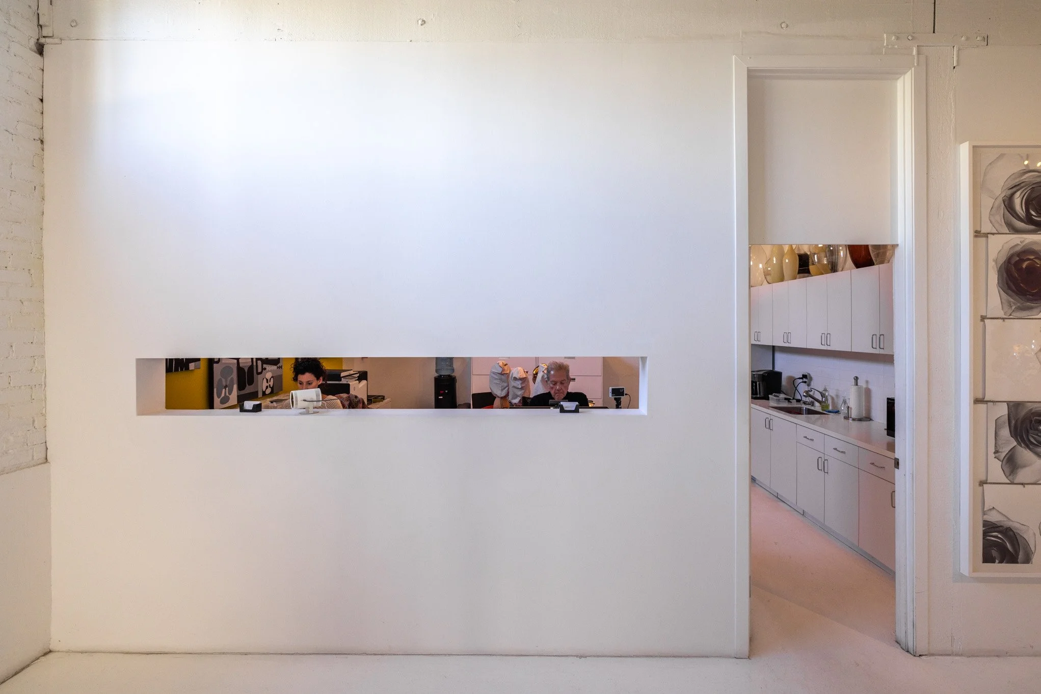 View through a rectangular horizontal window in a white wall, showing two people working in an office space with white cabinetry and kitchen appliances.