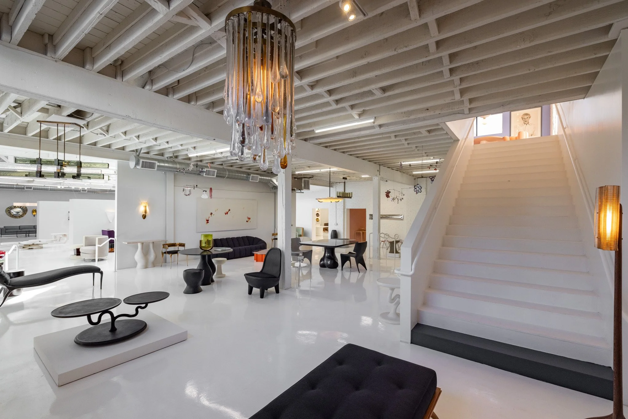 Modern interior of a spacious, minimalistic lounge or gallery with white floors and walls, showcasing contemporary furniture, a decorative chandelier, and a white staircase leading to an upper level.