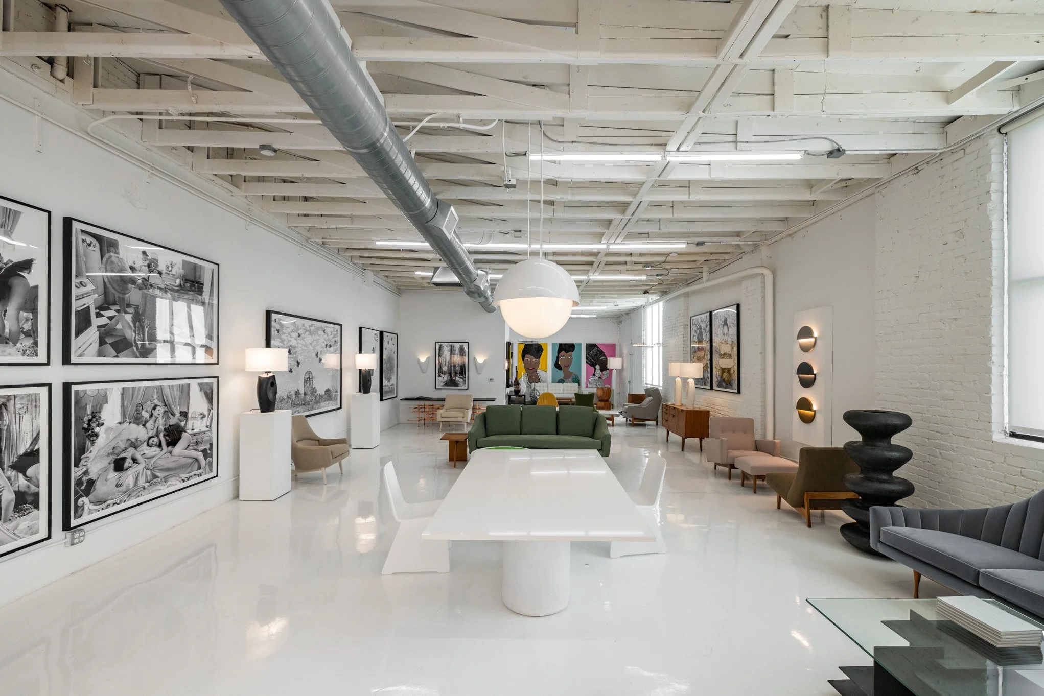 Modern art gallery with white walls, black and white photographs, colorful paintings, and eclectic furniture including sofas and chairs, illuminated by lamps and natural light.