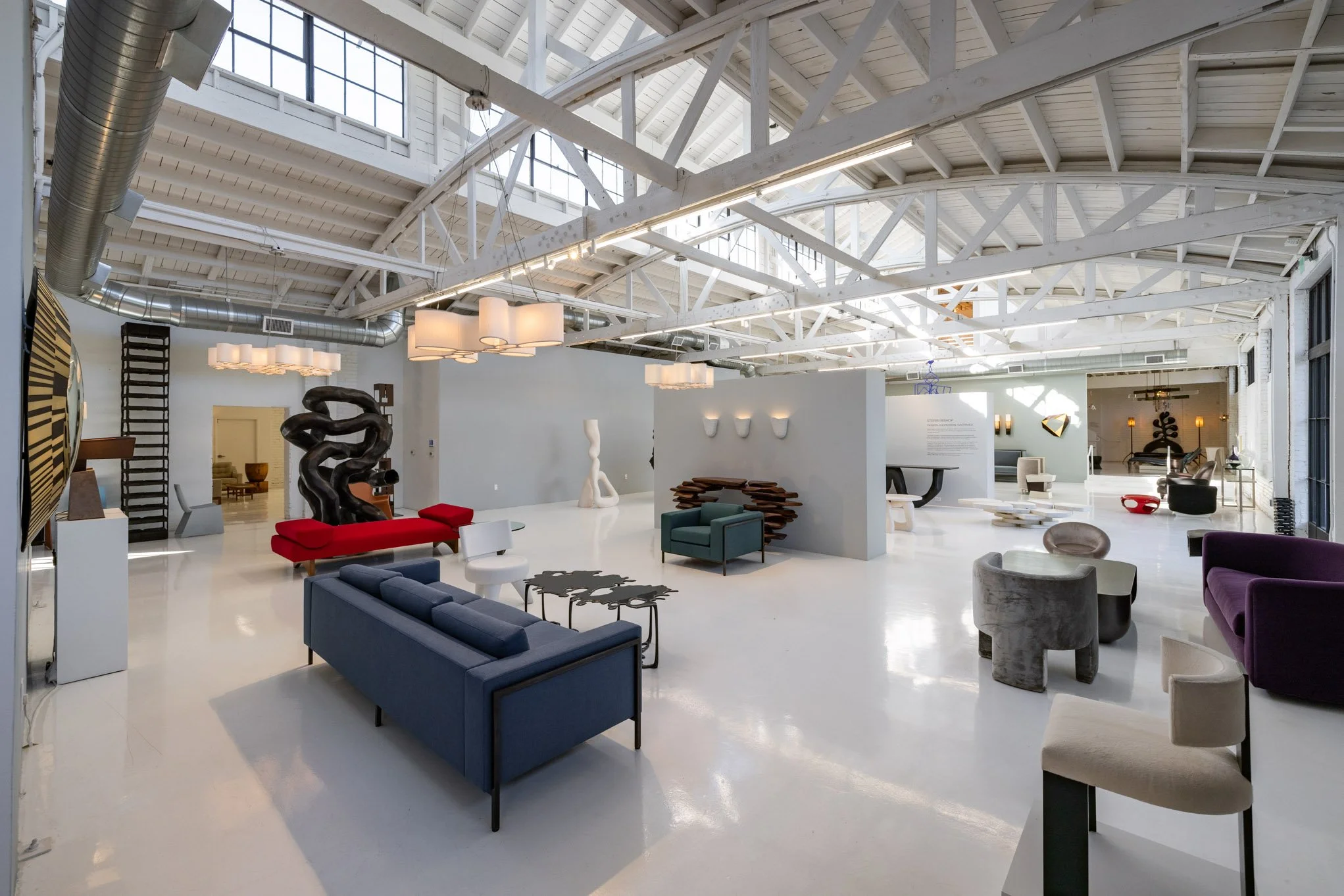 Interior of an art gallery or showroom with modern furniture and sculptures, high ceilings with exposed white beams, skylights, and large windows allowing natural light.