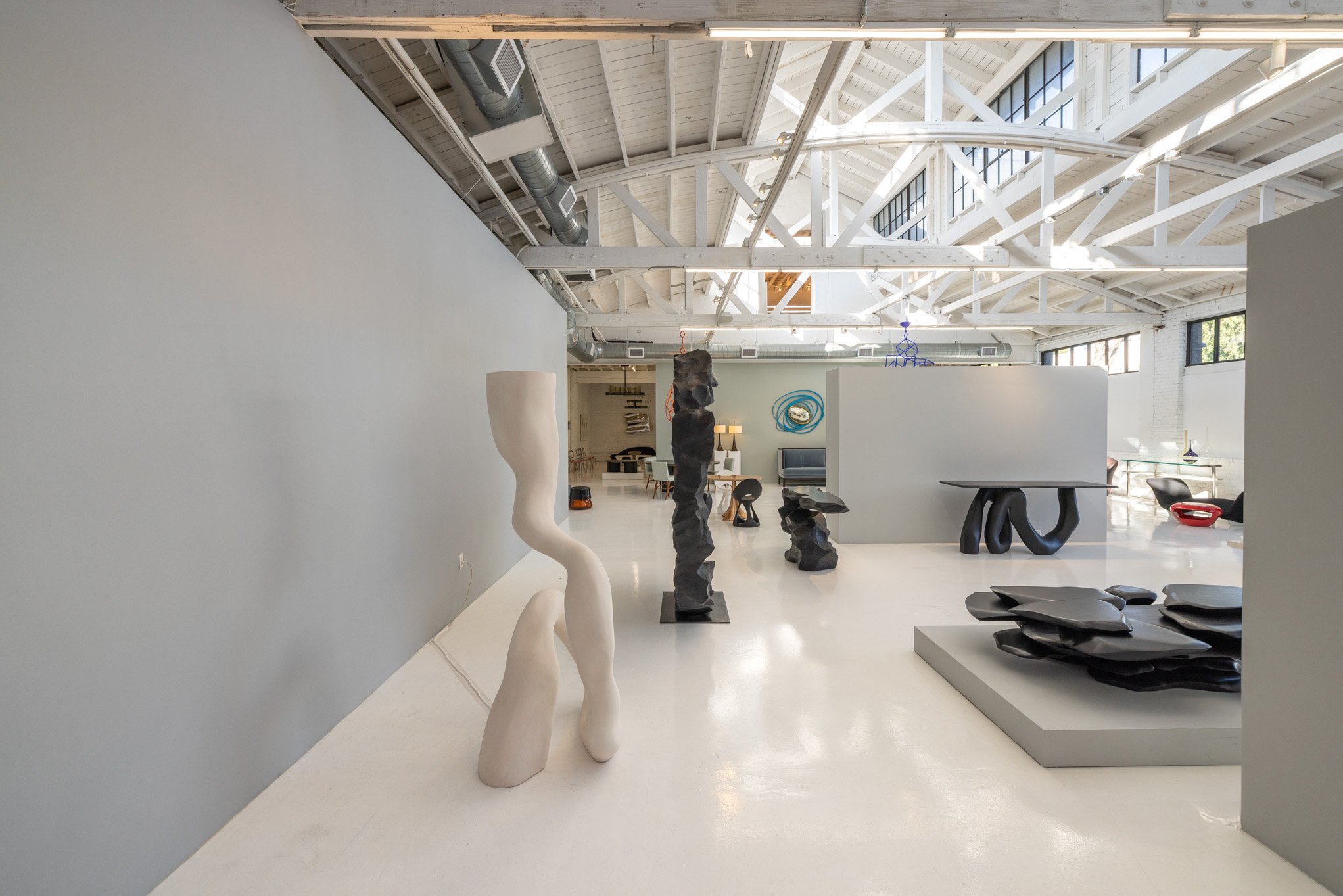 Interior of an art gallery with modern abstract sculptures and furniture, high ceiling with white exposed beams and windows, bright lighting.