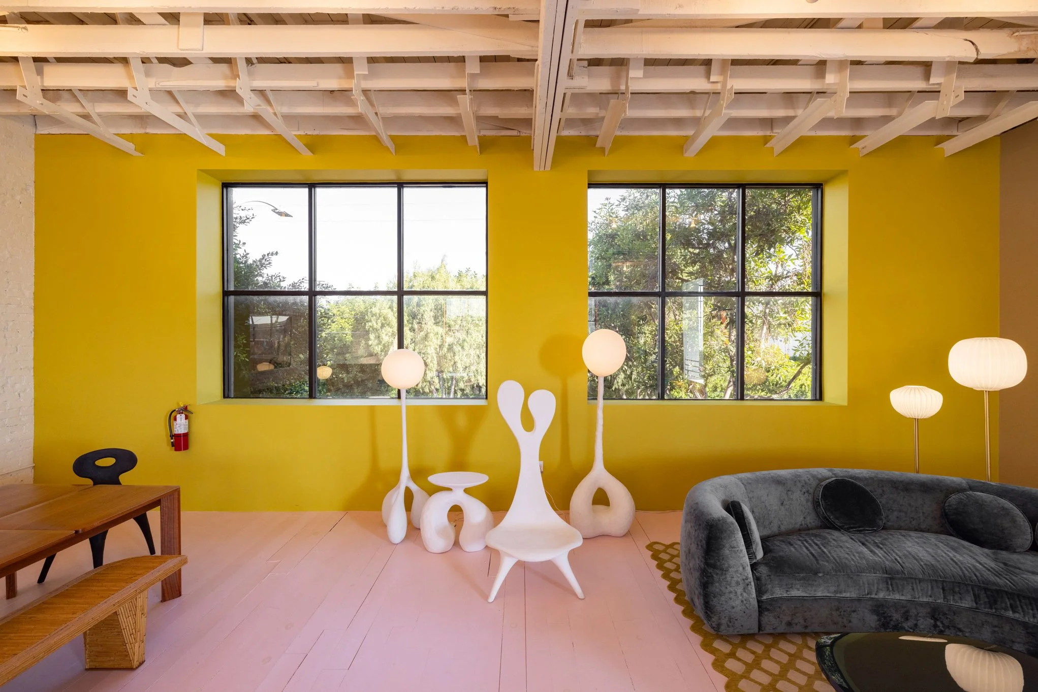 Living room with yellow accent wall, large windows, black window frames, artistic white floor lamps, a gray velvet sofa, and a wooden dining table.